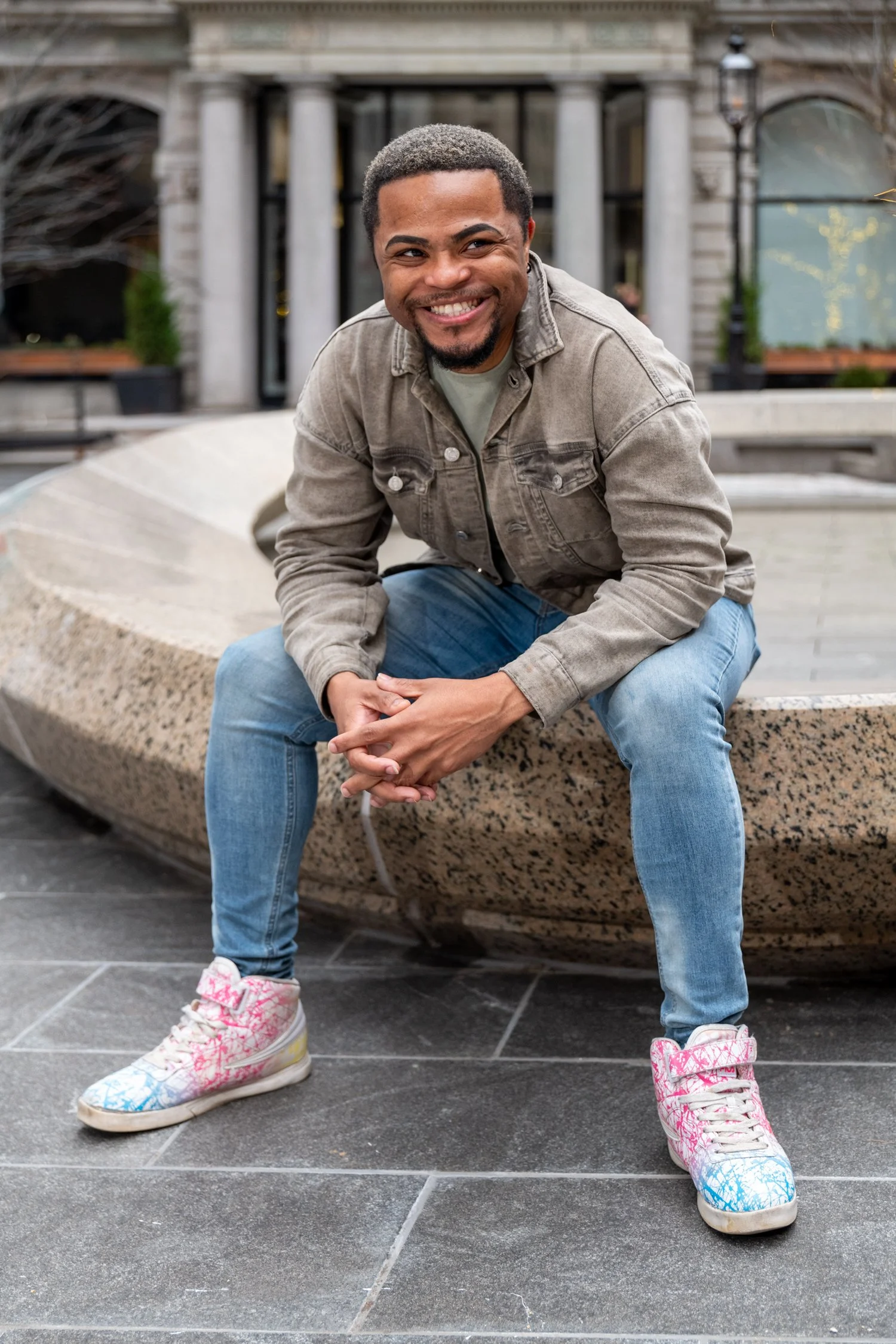 A smiling man with short curly hair and a beard, wearing a tan denim jacket, light blue jeans, and white sneakers with pink and blue spray paint designs, is squatting on a stone ledge in an urban outdoor setting.