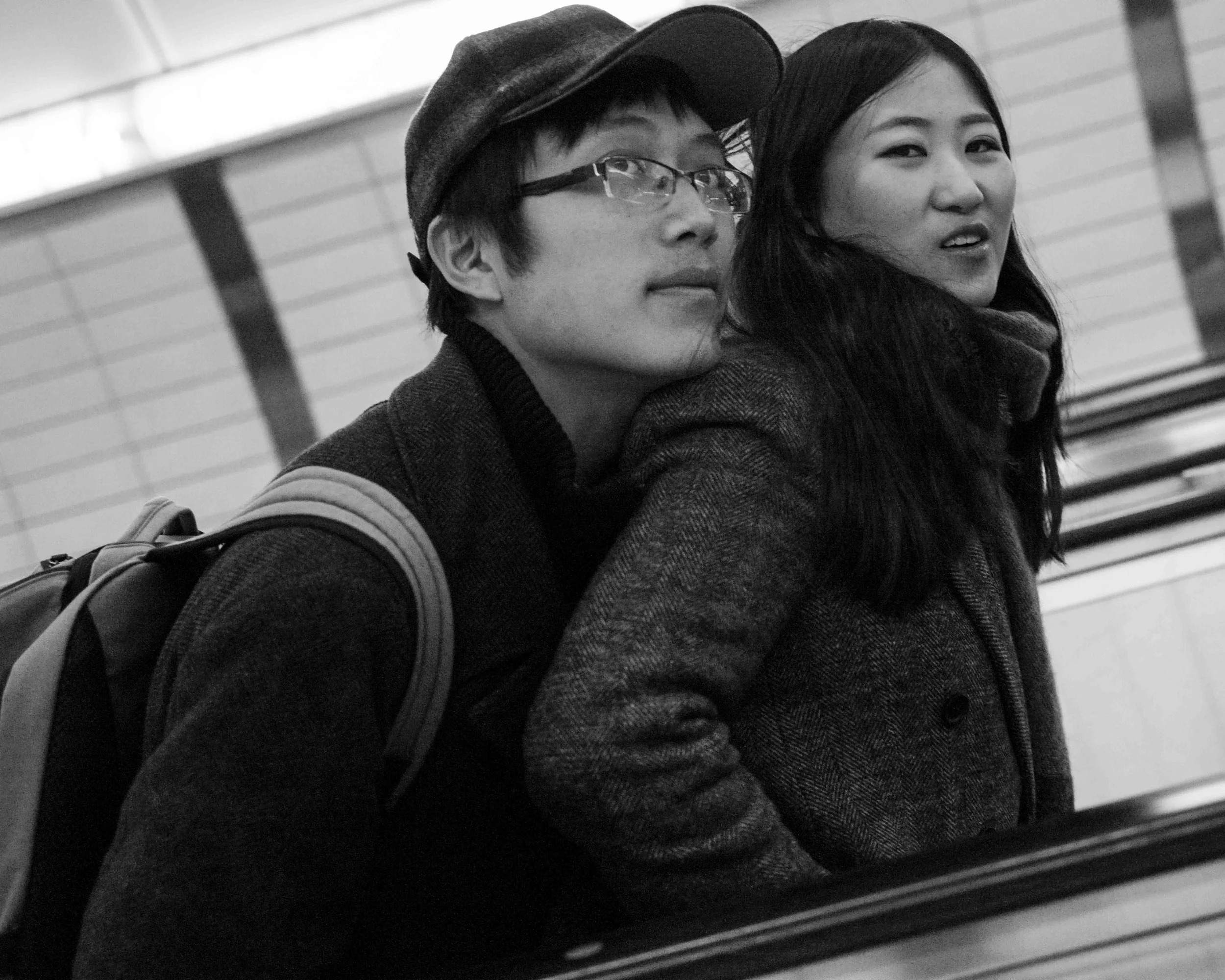 Black and white photo of a young man wearing glasses and a cap, and a young woman with long hair and a coat, sitting or standing close together.