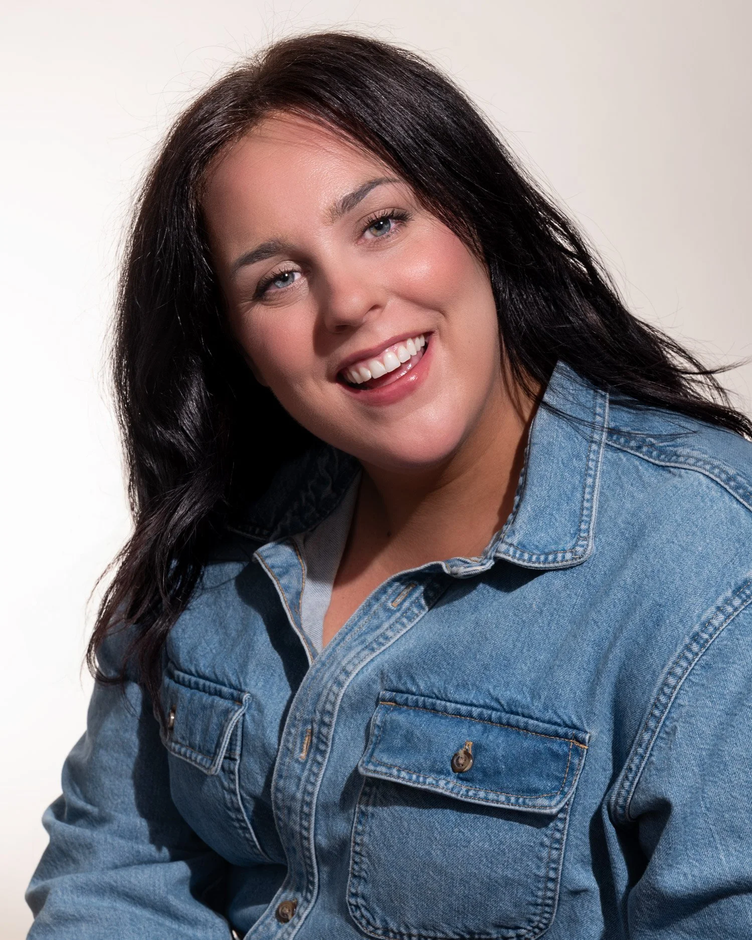 Close-up of a smiling woman with dark hair wearing a denim jacket against a white background.