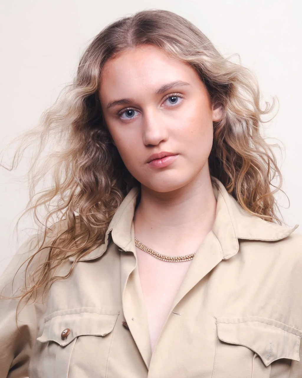 A young woman with long, wavy blonde hair and blue eyes, wearing a beige button-up shirt with chest pockets and a gold chain necklace, looking directly at the camera against a plain light background.
