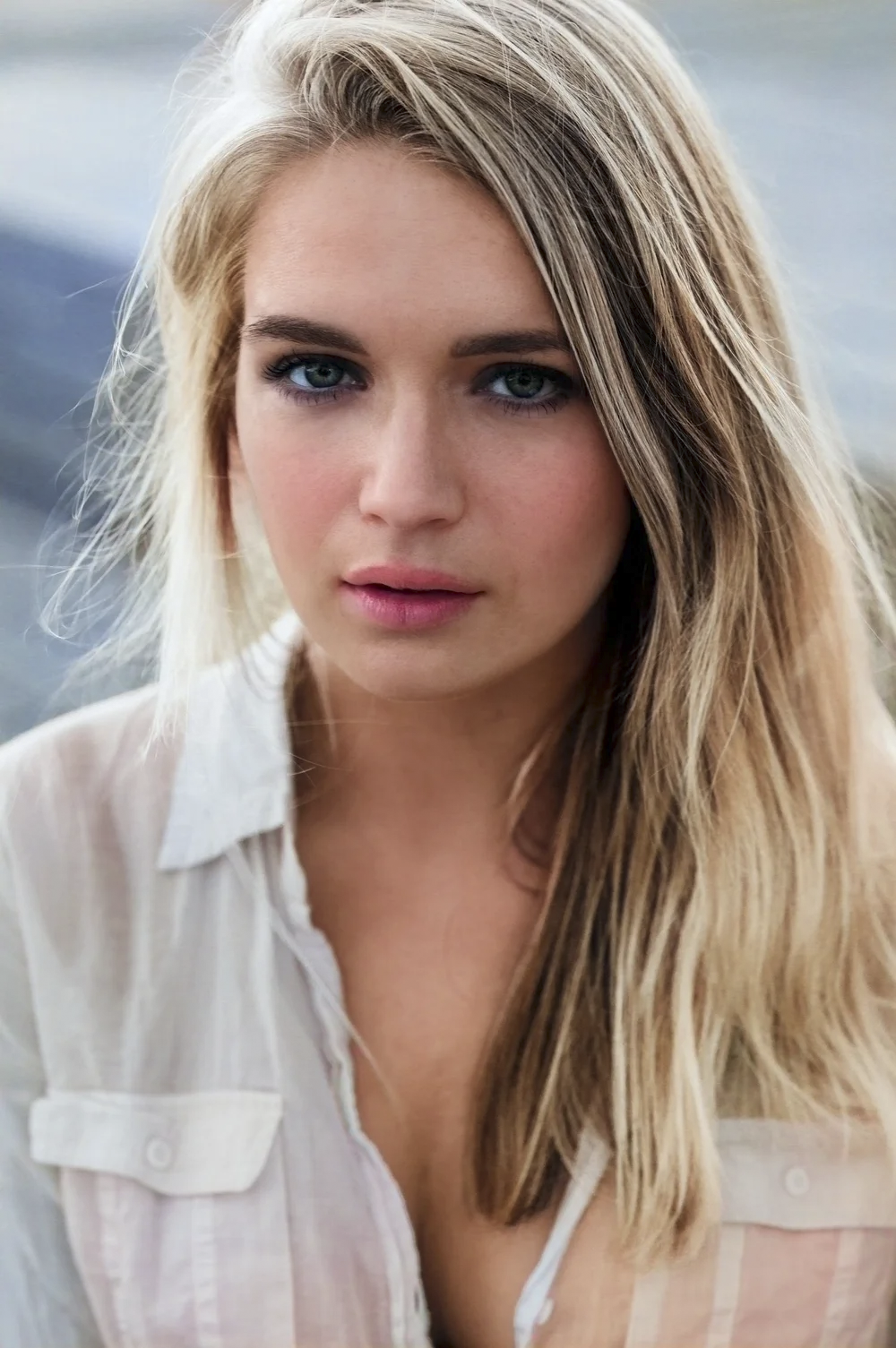 Close-up of a young woman with long blonde hair and blue eyes, wearing a white shirt, standing outdoors during daytime.
