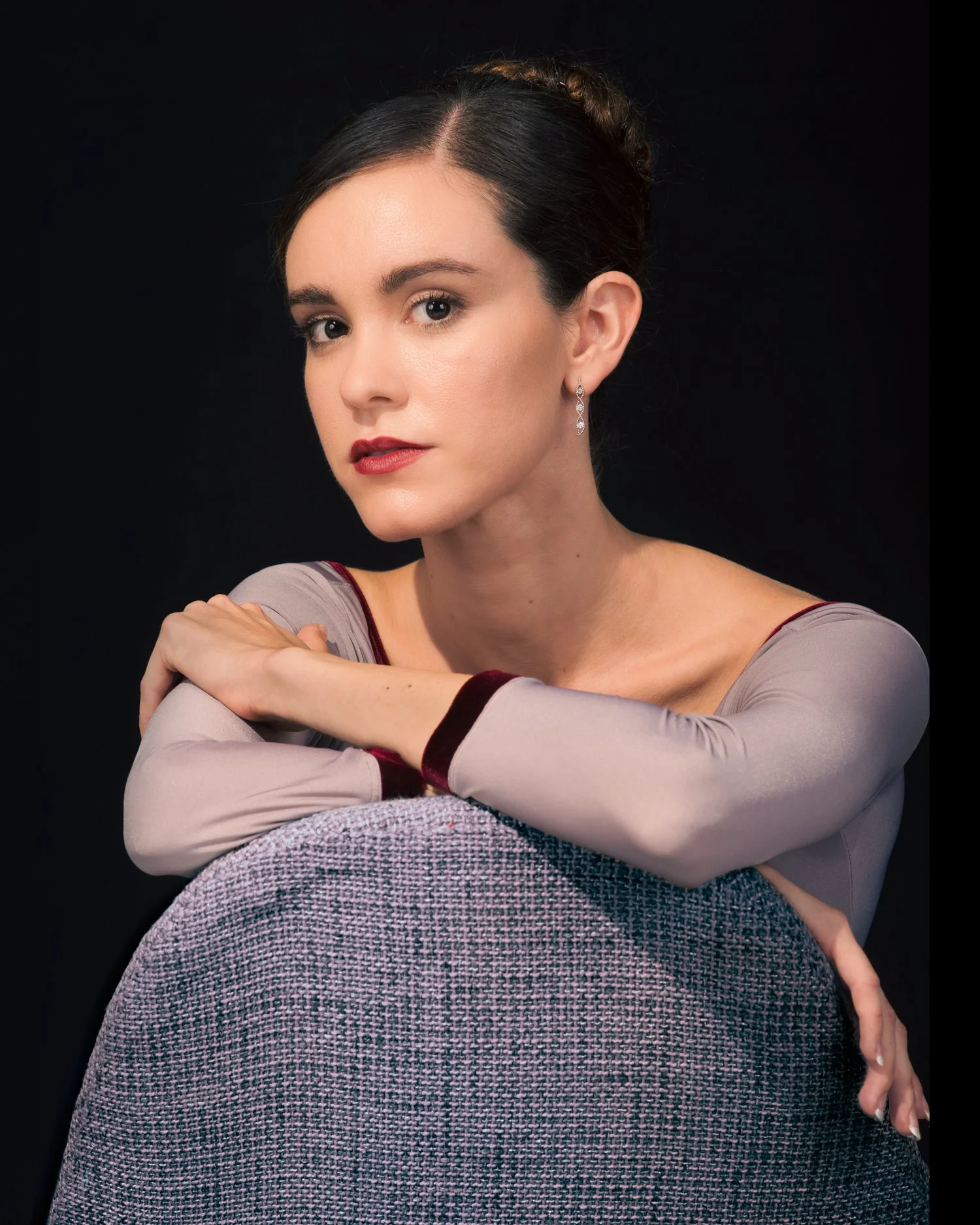 Close-up portrait of a woman with dark hair, fair skin, and red lipstick, wearing earrings and a gray top with maroon accents, against a black background.