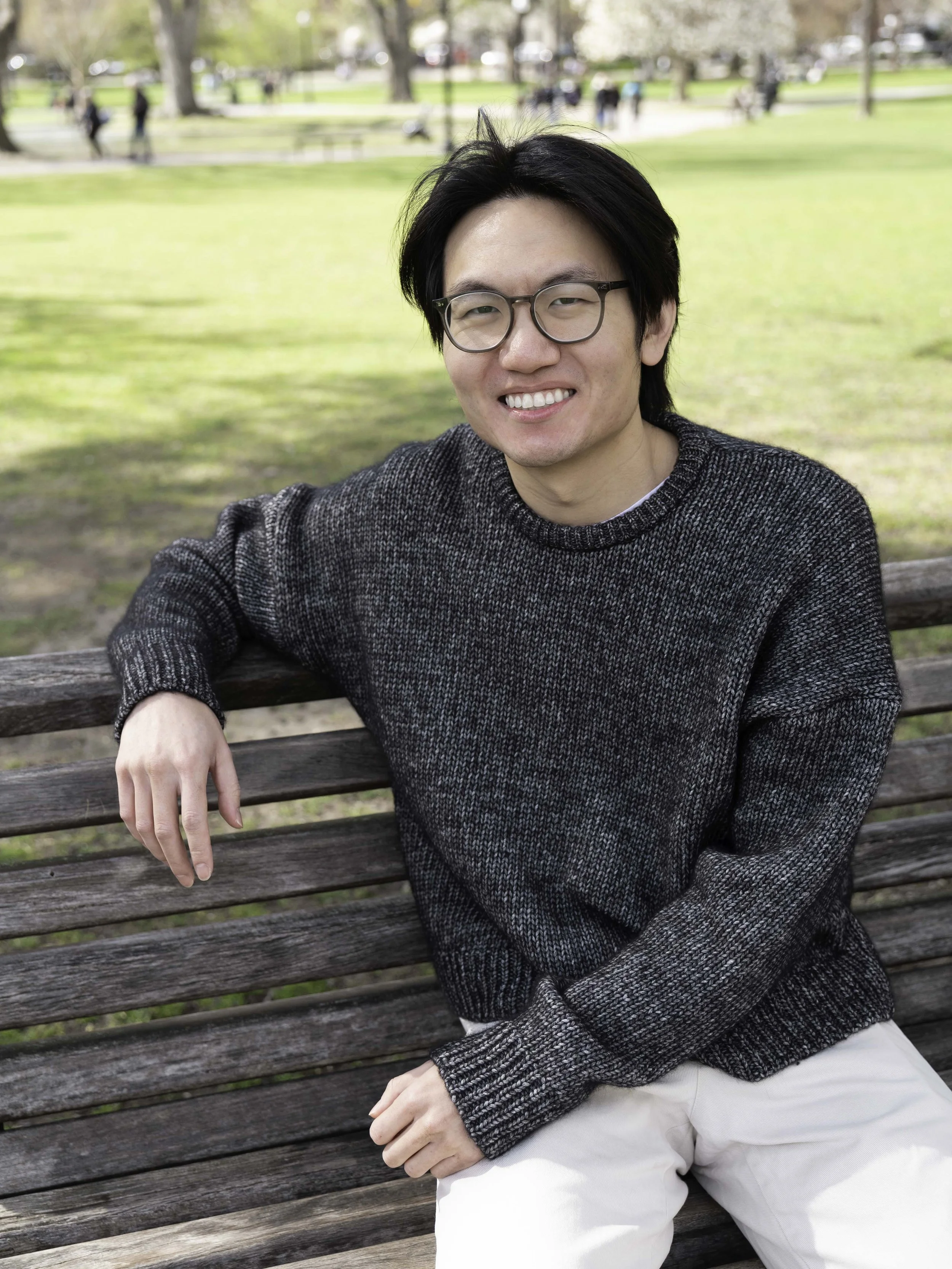 Young asian man in a gray sweater and white pants seated on a park bench in sunlight with green lawn behind him.