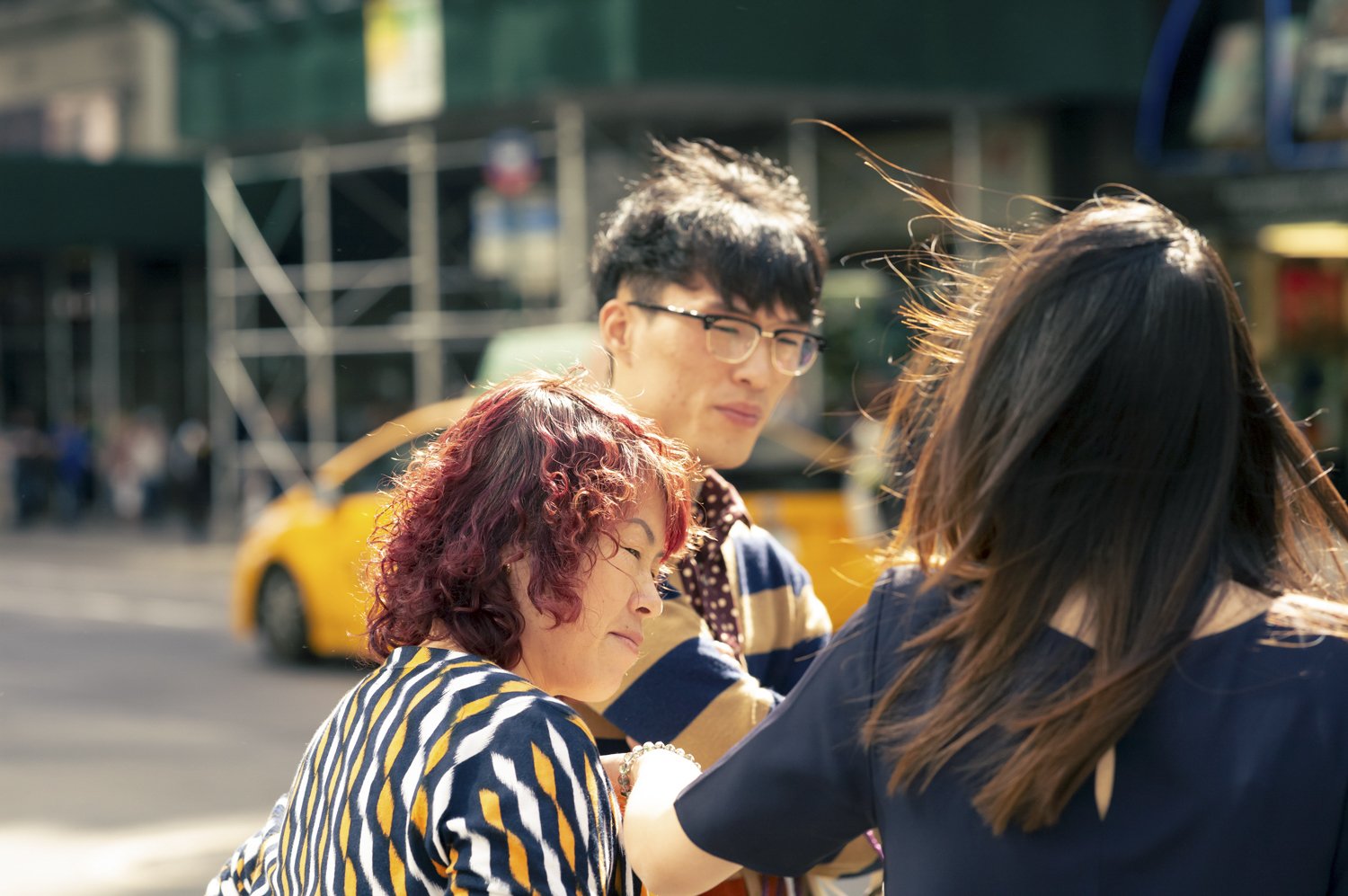 Three people, two women and one man, standing closely together outdoors in what appears to be a city street, with a yellow taxi cab and urban buildings in the background. The woman with curly red hair and striped shirt is smiling, the man with glasse