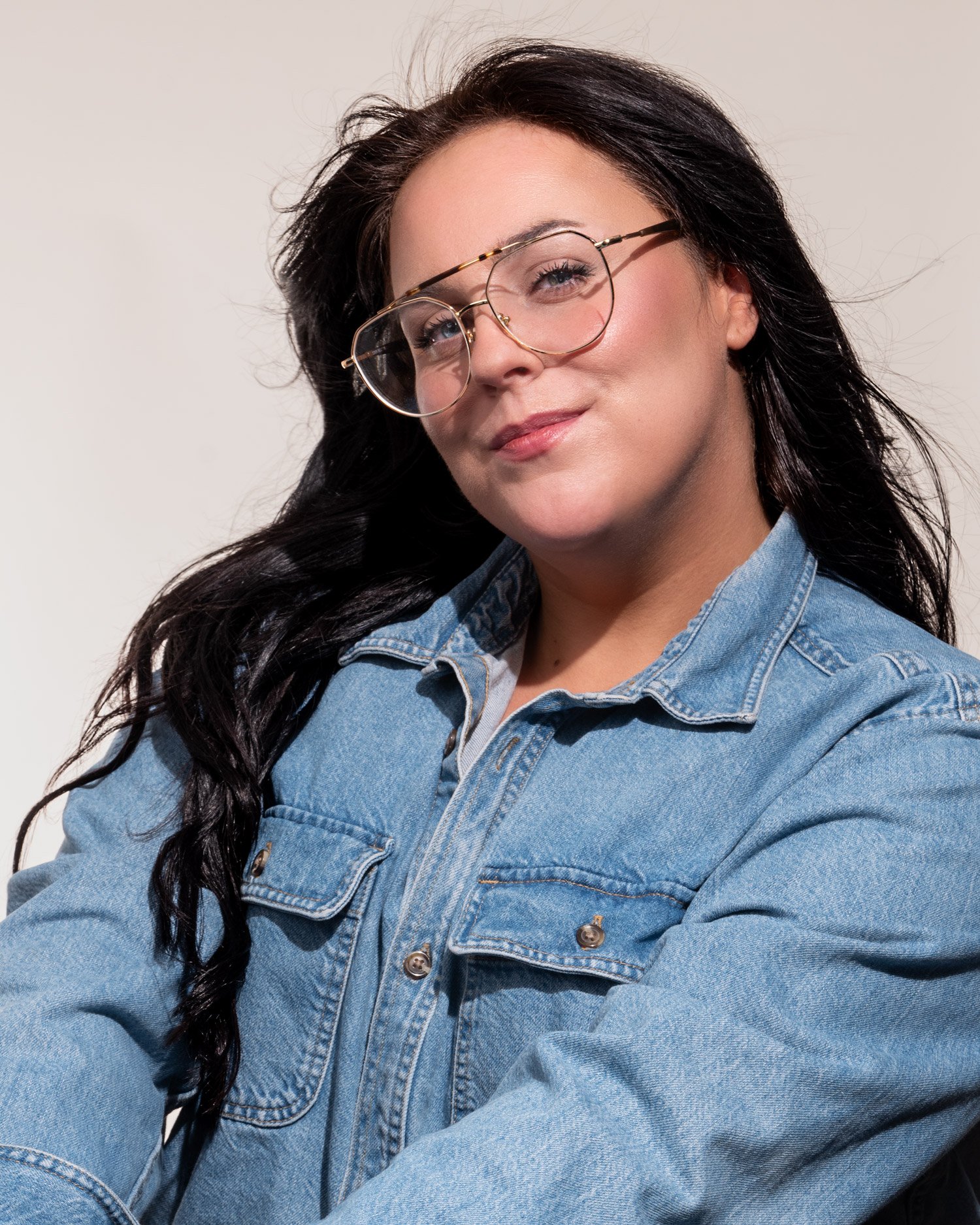 A woman with long dark hair, wearing glasses and a denim jacket, smiling softly against a plain background.