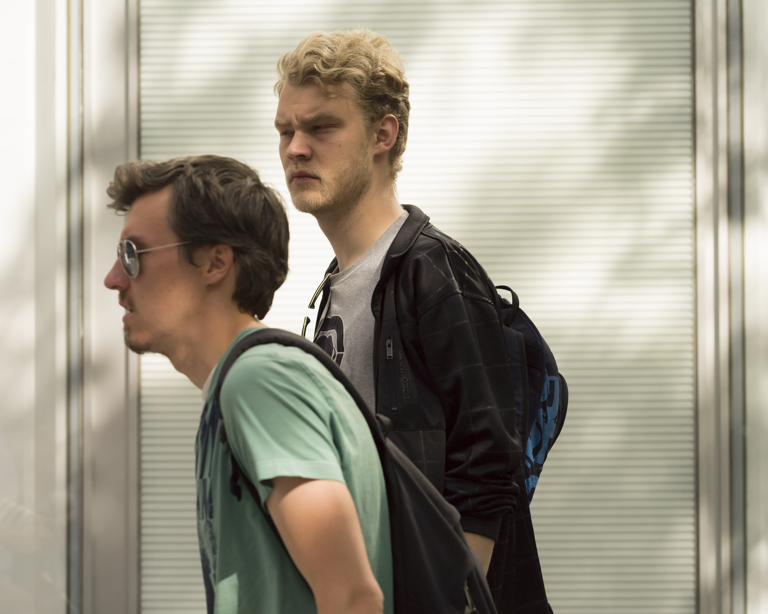 Two young men standing inside an elevator with metallic walls, one with curly blonde hair wearing a gray shirt and black jacket, the other with short dark hair and sunglasses, wearing a green T-shirt and carrying a backpack.