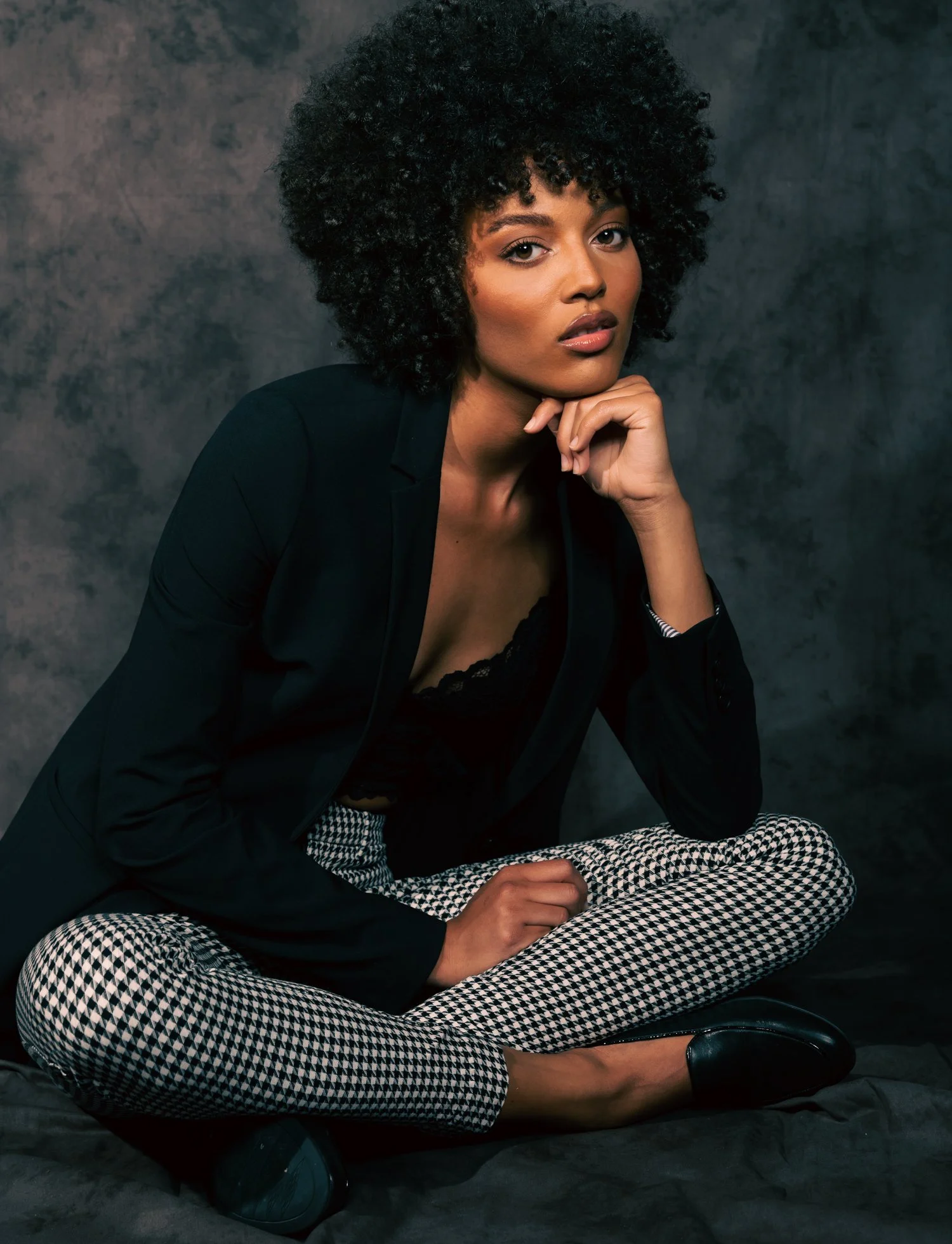 Model seated cross-legged on a floor against a bluish-gray fabric backdrop, with her chin rested against her hand. She has voluminous black, curly hair with tan skin and well-defined facial structure and wears a low-cut black top with black and white