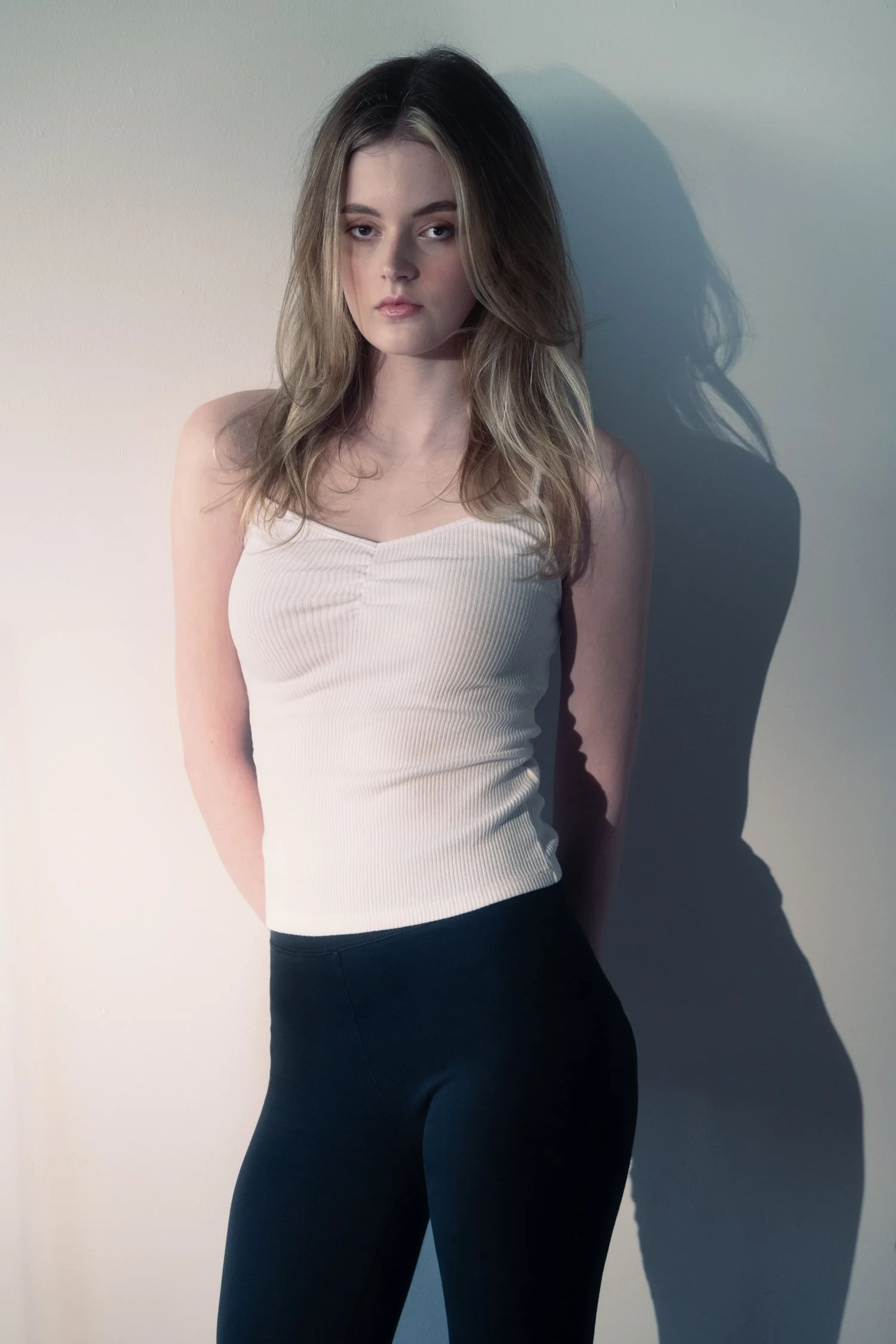 Young woman poses for her model card digitals in a white tank top and black leggings in a studio setting with dynamic shadows.