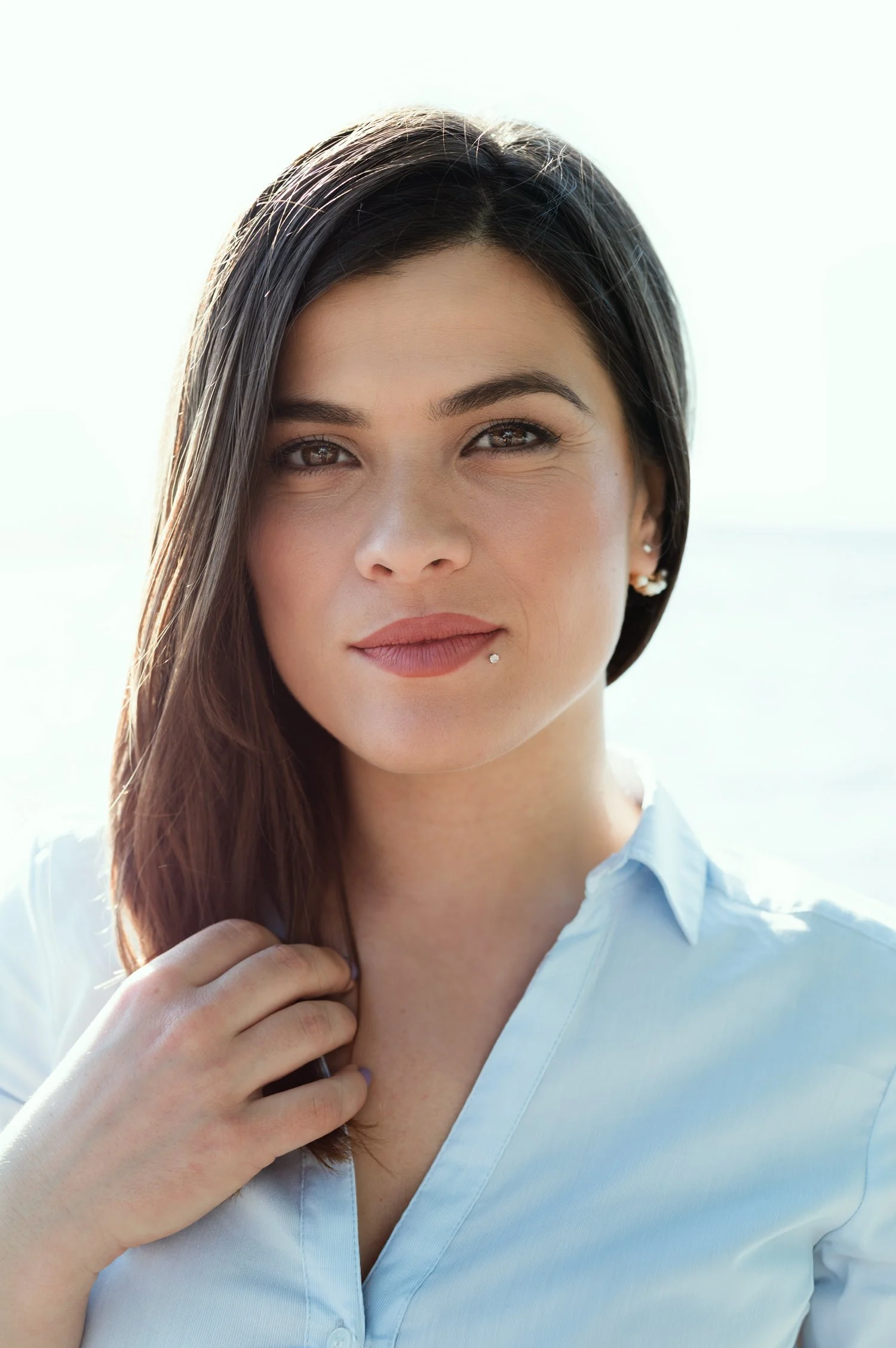 Natural outdoor daylight head and shoulders portrait of a young woman with long, dark hair, wearing a light blue shirt. Her right hand is stroking her hair.
