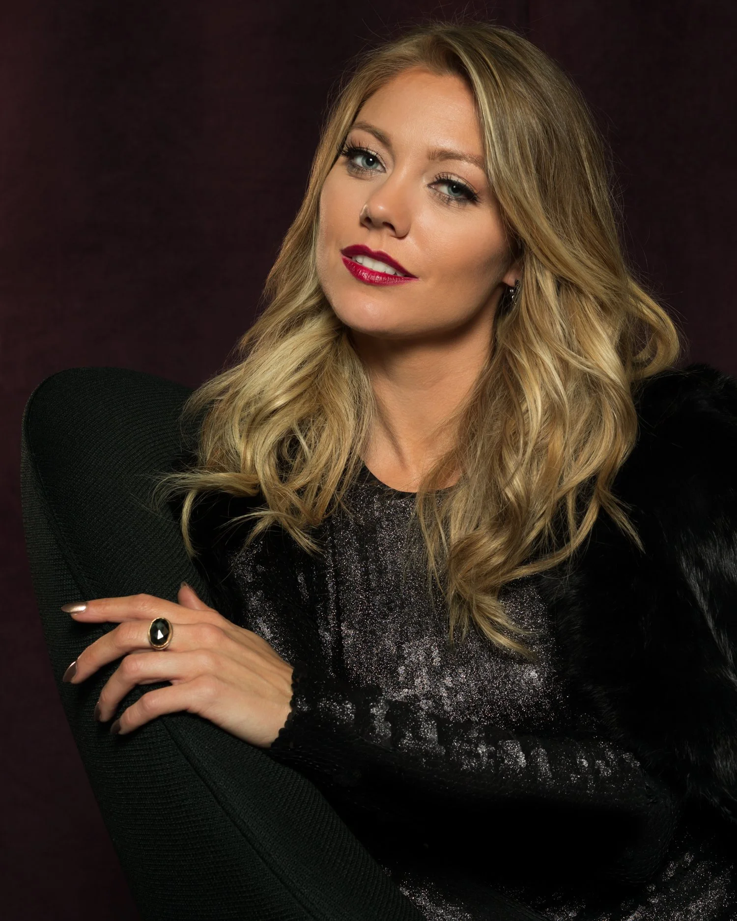 A woman with blonde wavy hair, wearing a black top with glitter, sitting on a black chair with a dark background.