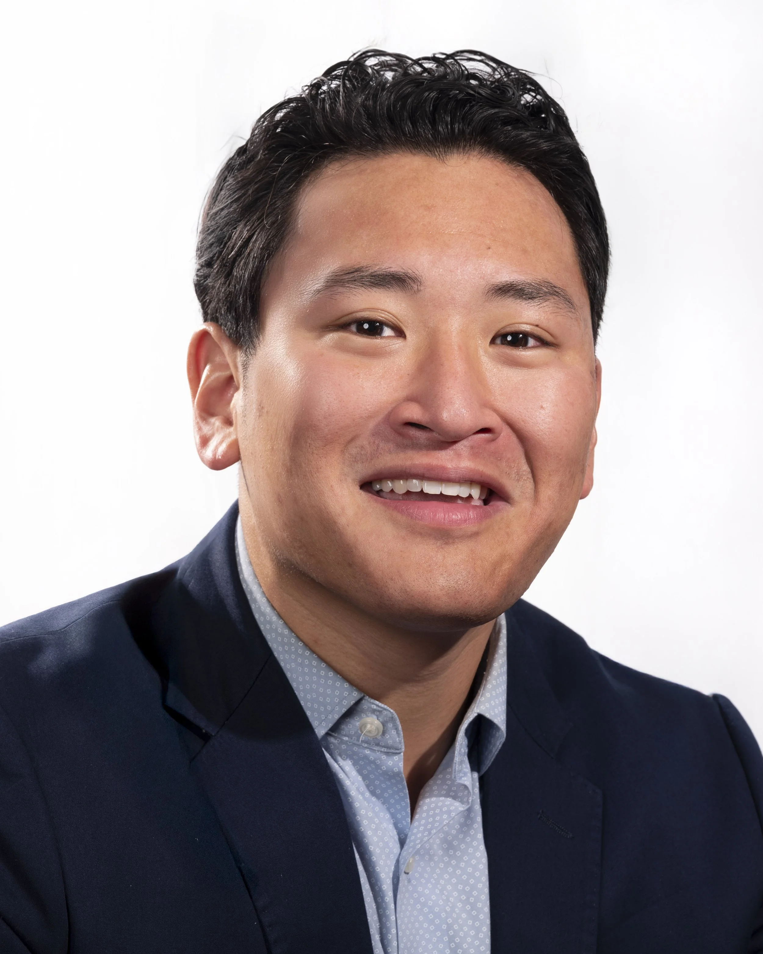 Headshot of smiling young Asian man wearing a dark blue blazer and light blue shirt against a white backdrop.