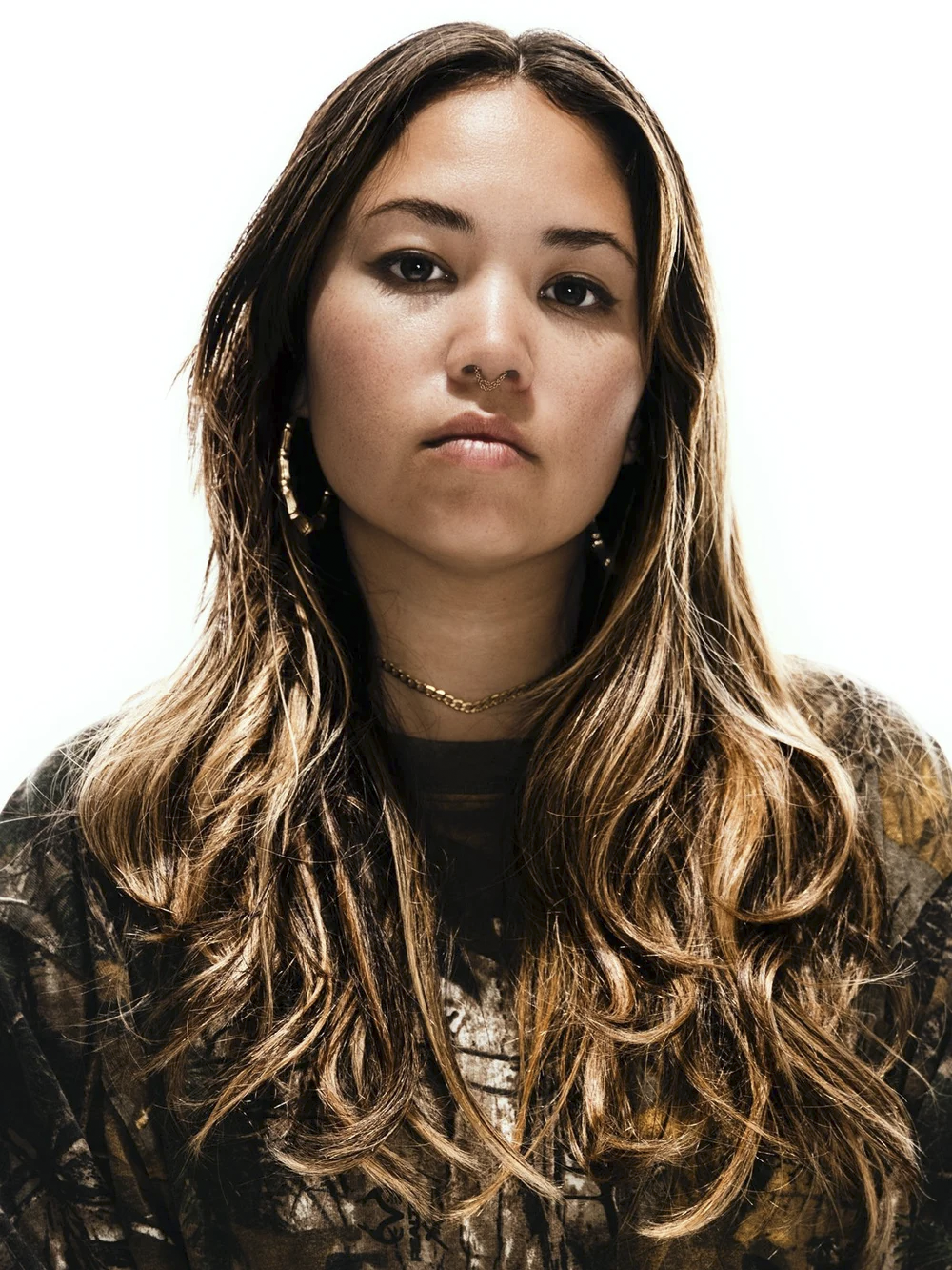 Portrait of a young woman with long, wavy brown hair, wearing hoop earrings, a septum piercing, a necklace, and a black camouflage-patterned shirt, against a plain white background.