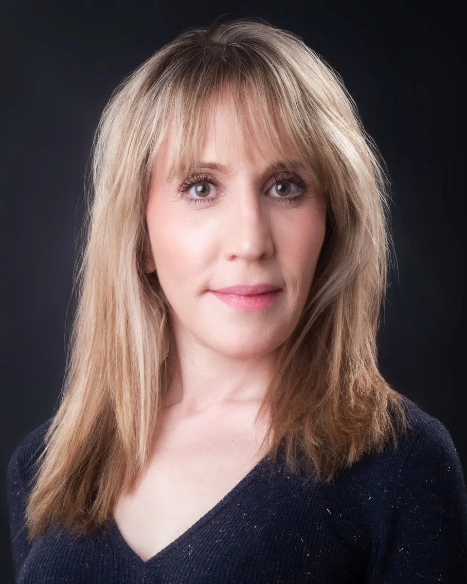 Headshot of a woman with straight, shoulder-length blonde hair, wearing a dark blue top featuring sparkly bits, photographed on a dark background.