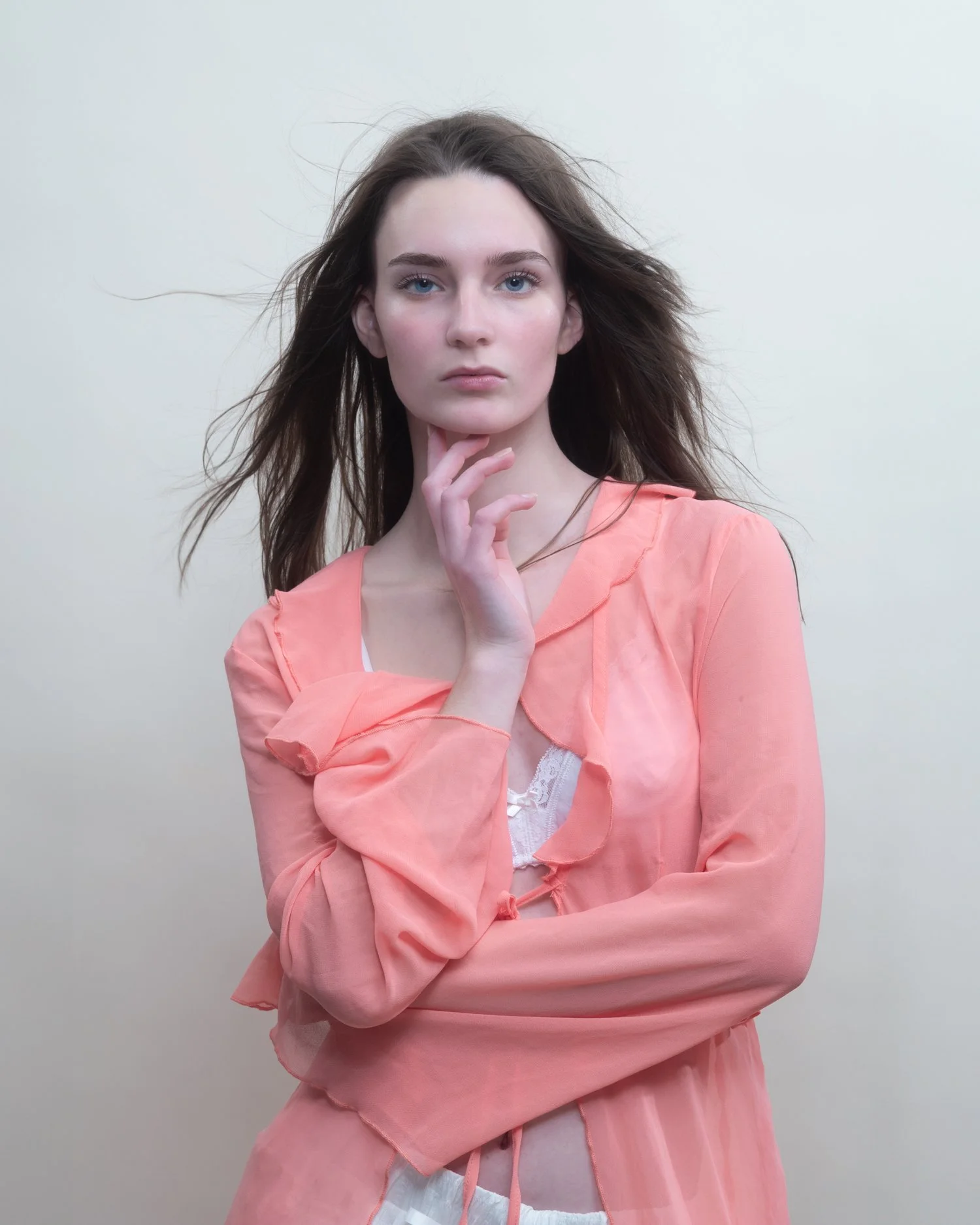 A young woman with long, wavy brown hair and blue eyes, wearing a light pink, semi-sheer blouse and white undergarments, standing against a plain, light-colored background, with her left hand resting on her chin.