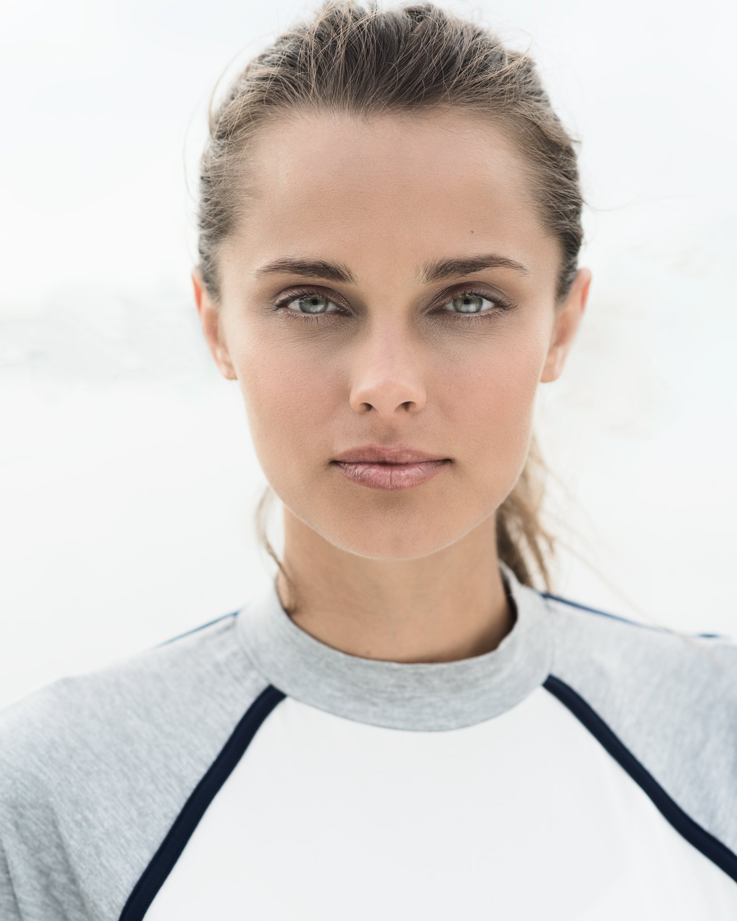 Closeup portrait of young woman with pulled-back light brown hair, wearing fitness wear. Photographed outdoors in natural light, the background is blown-out white haze.