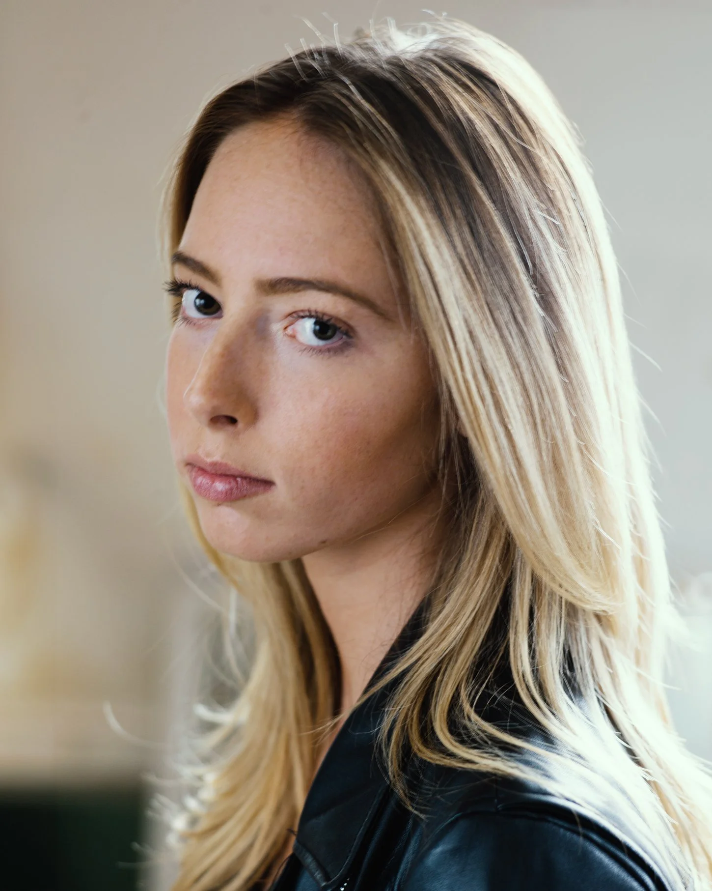 Close-up of a young woman with blonde hair and blue eyes wearing a black leather jacket, looking over her shoulder at the camera.