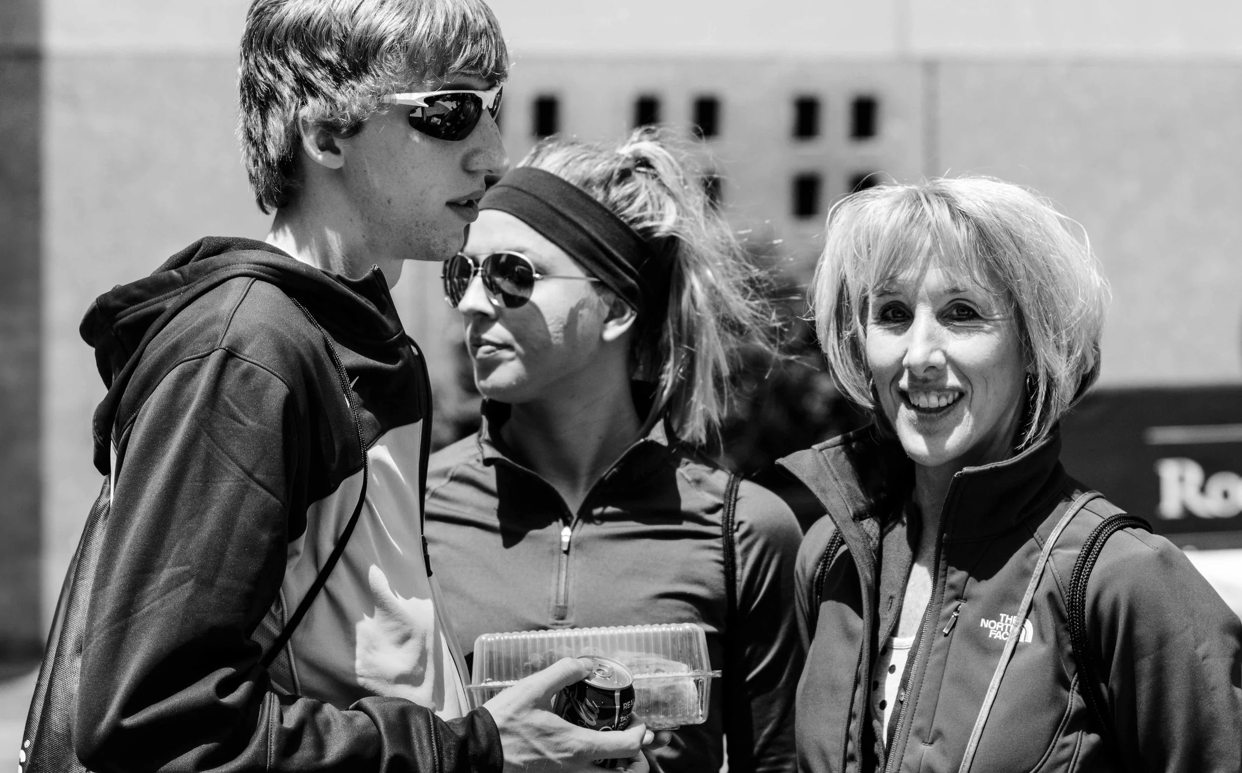 Four women standing in a group outdoors, engaged in conversation, with buildings in the background, all wearing jackets and sunglasses.