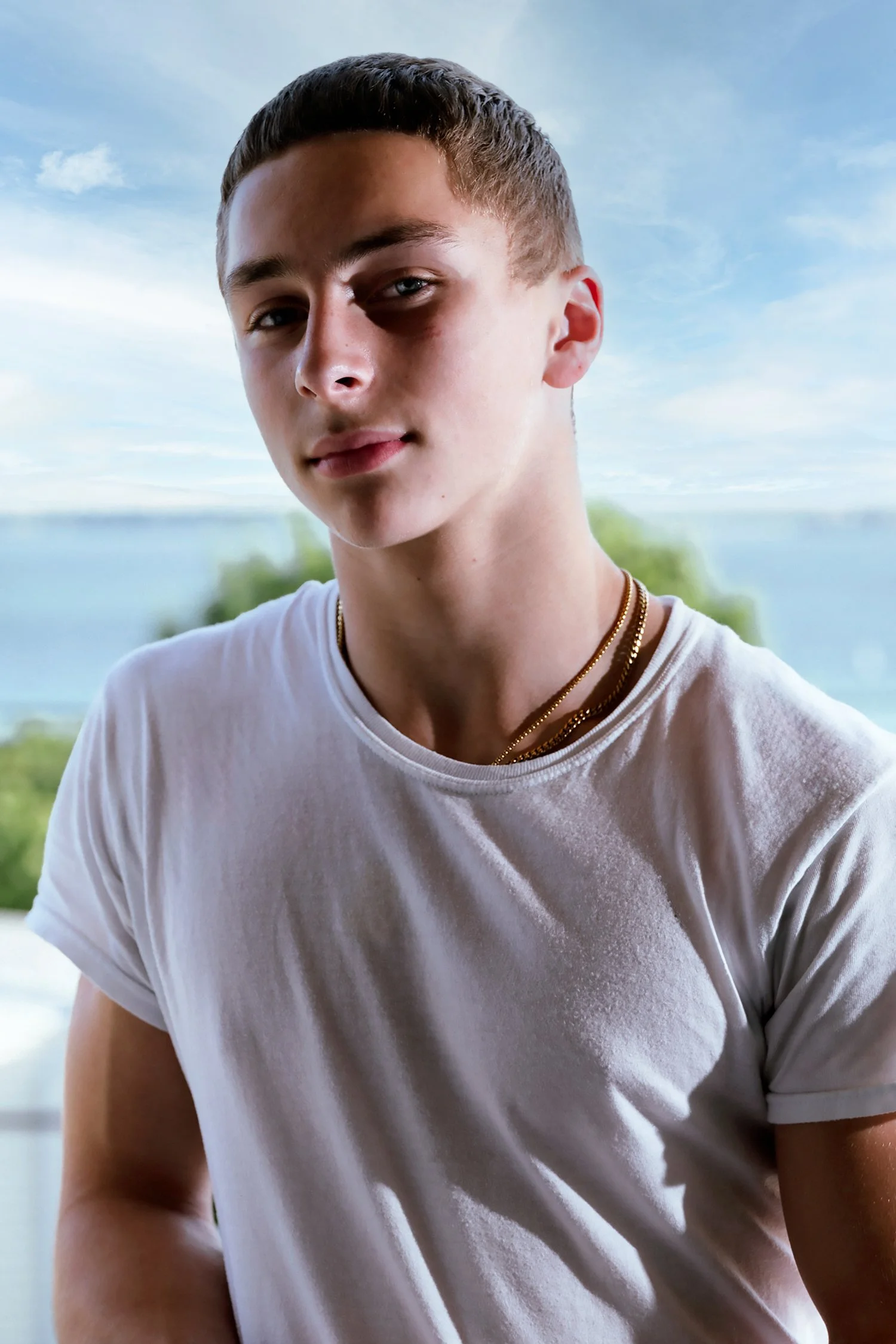 Young man with short brown hair wearing a white T-shirt, multiple gold necklaces, and bracelets, standing outdoors near a body of water with trees and a house in the background.