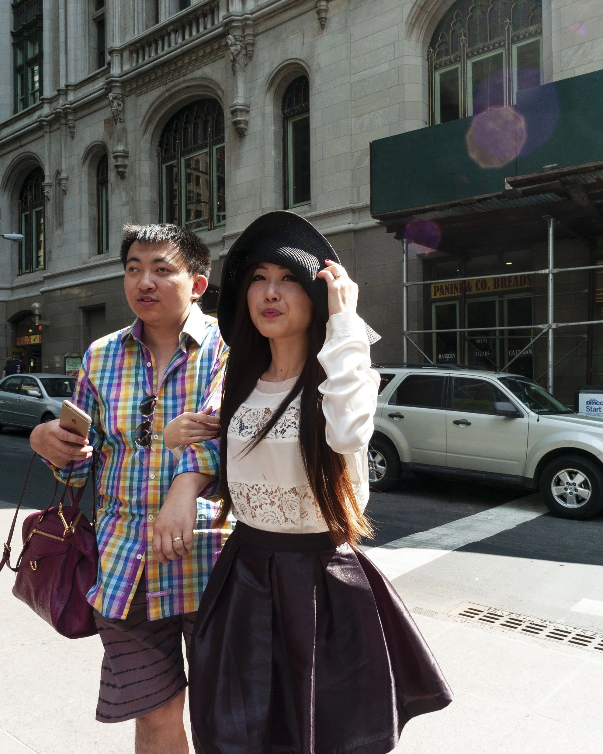 Two people walking on a city street in front of a historic building with ornate windows. The woman is wearing a black hat, white lace top, and dark skirt, and is holding her hat with one hand. The man is wearing a colorful plaid shirt and holding a s