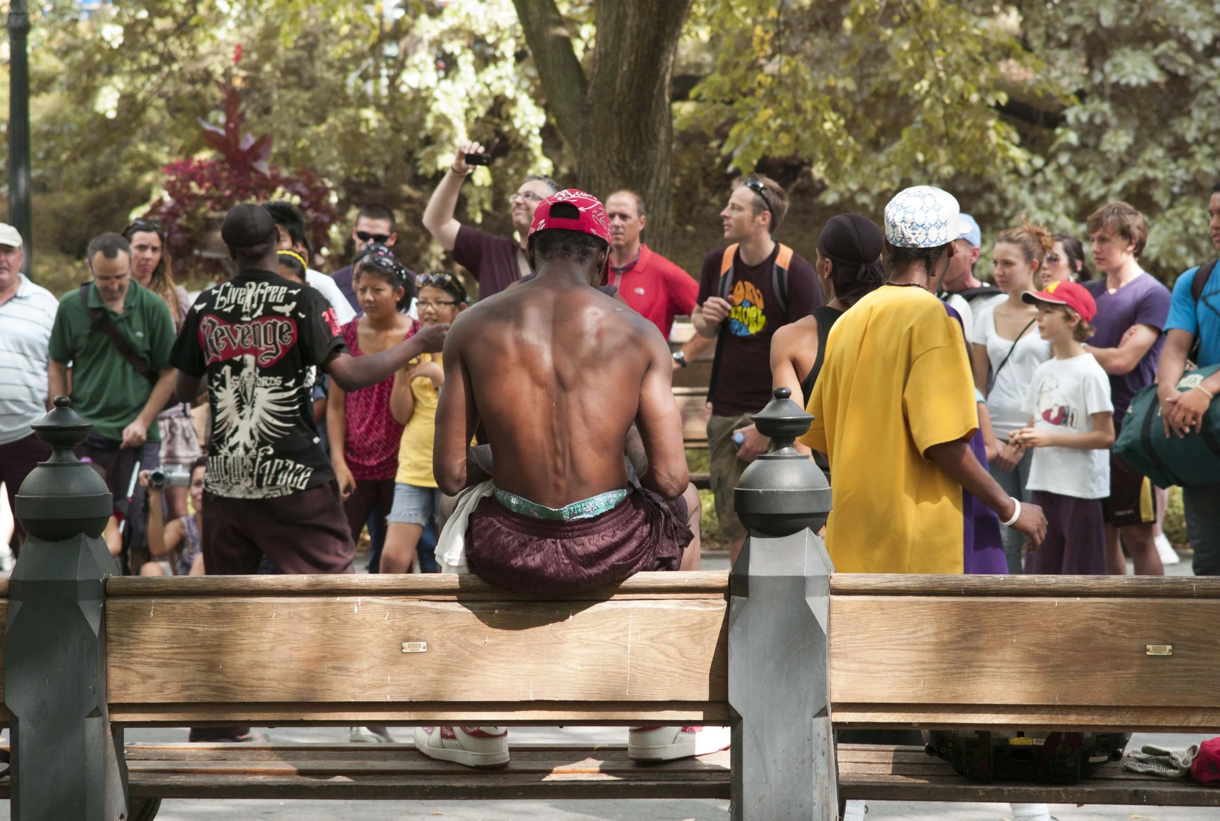 A shirtless man with dark skin and a red cap, sitting with his back to the camera on a wooden bench in a park surrounded by a group of diverse people. The group includes men, women, and children of various ages and ethnicities, some wearing backpacks