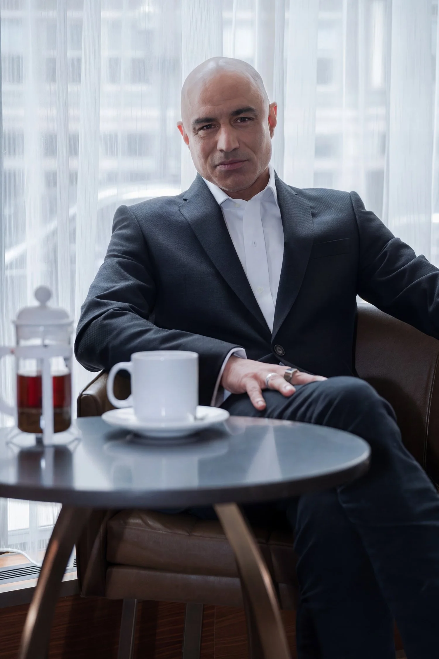 A bald man in a business suit sits backlit in front of a large hotel window with a coffee pot, cup, and saucer on a table in front of him.