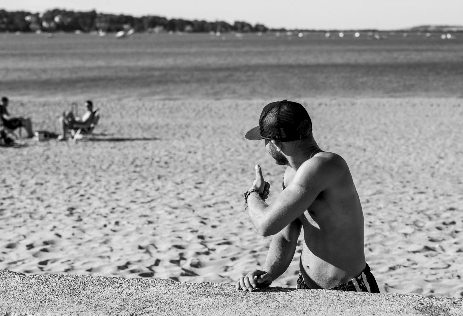 A shirtless man with a beard and cap making a gesture with his hand on a beach with a few people sitting in the background.