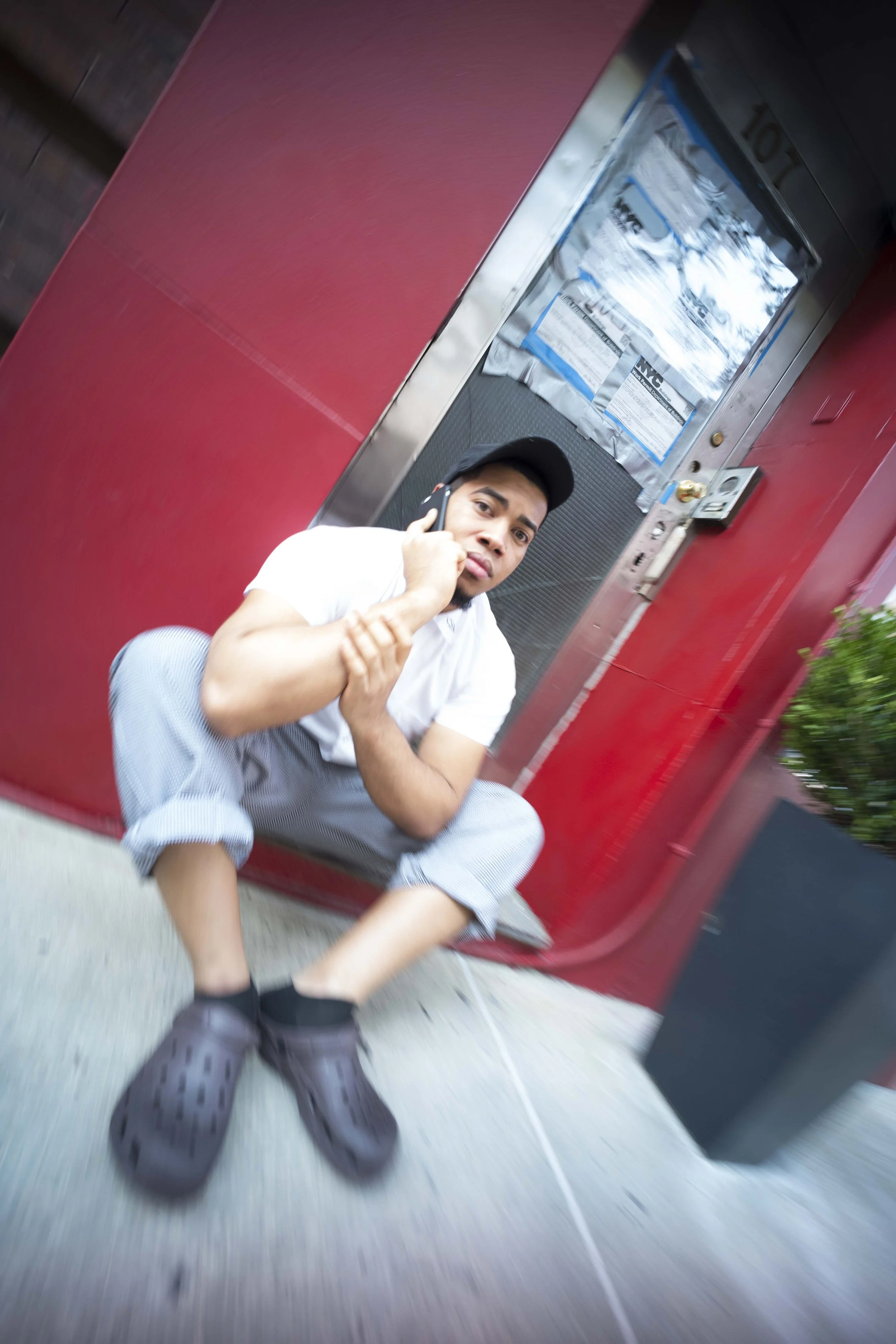 A young man in casual clothing and crocs shoe, squatting on a sidewalk, talking on a cell phone with a expression of concentration. He is in front of a red door with notices taped on it.