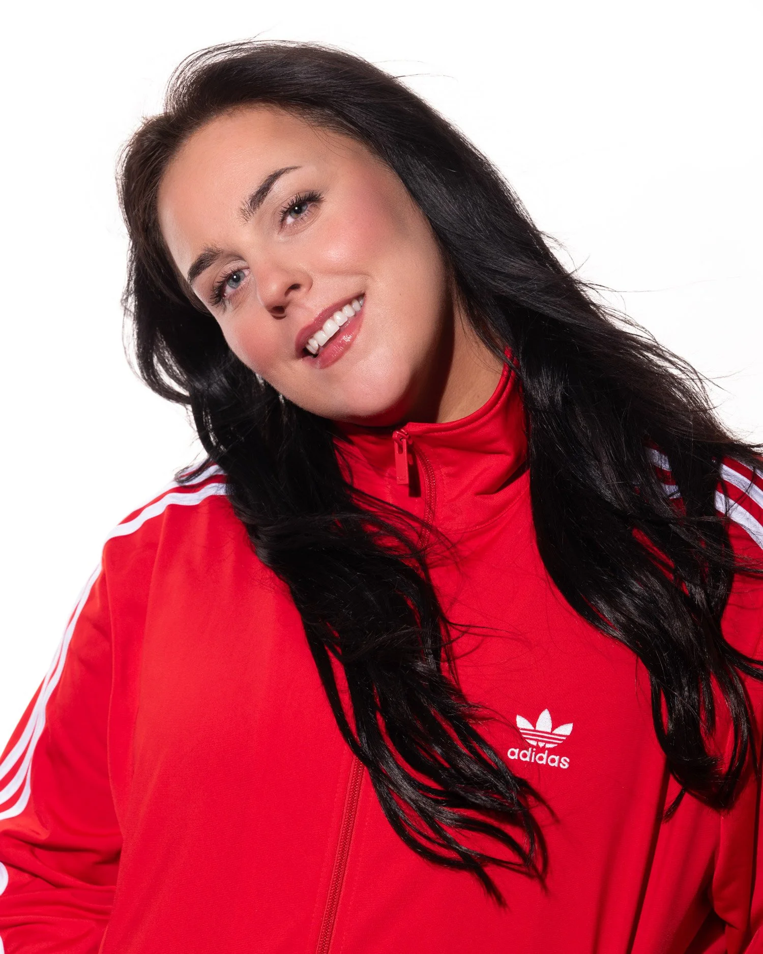 A woman with long black hair, blue eyes, and a fair complexion wearing a red Adidas jacket.