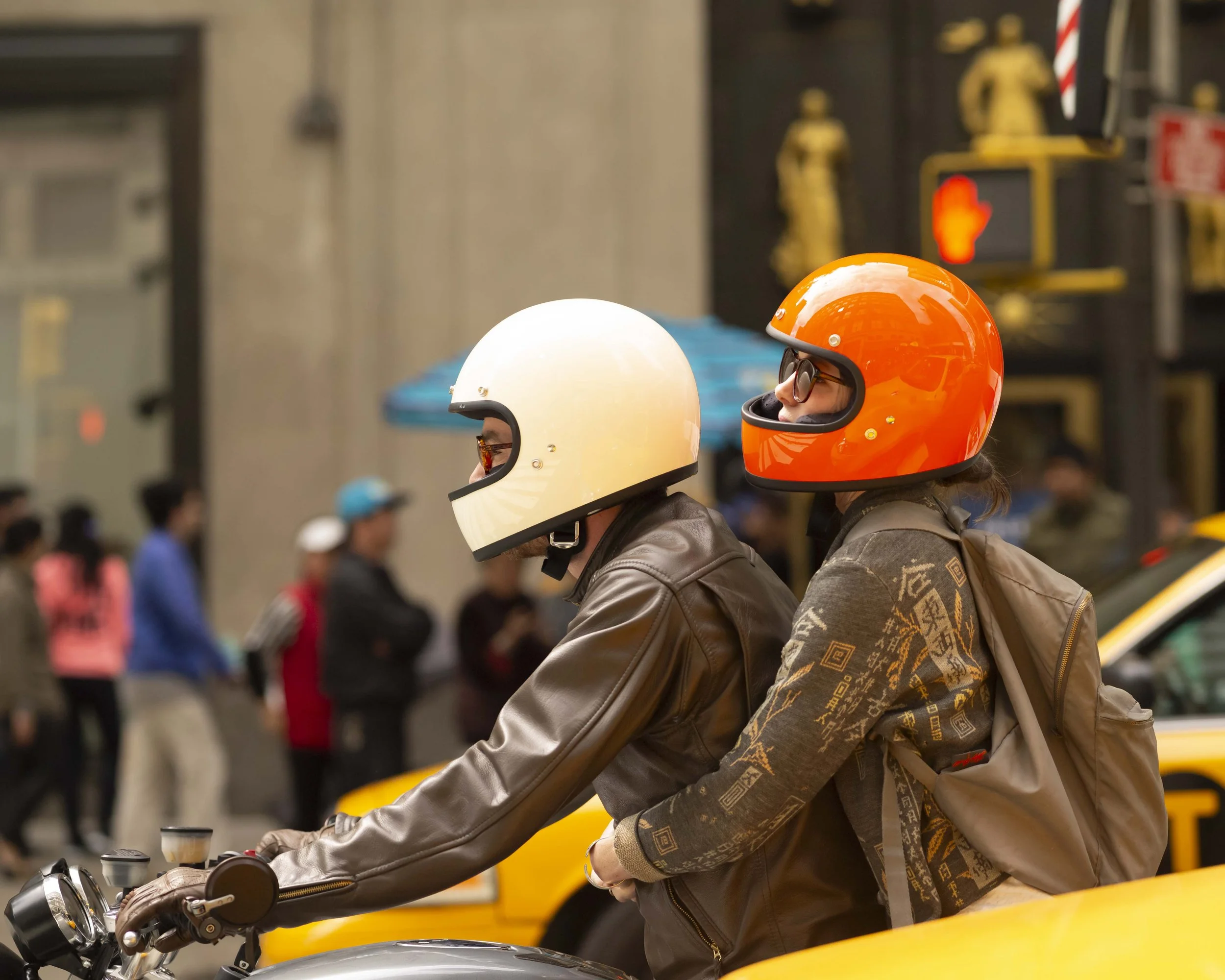 Two people wearing motorcycle helmets riding on a motorcycle in an urban area, with a crowd of pedestrians and a yellow taxi in the background.