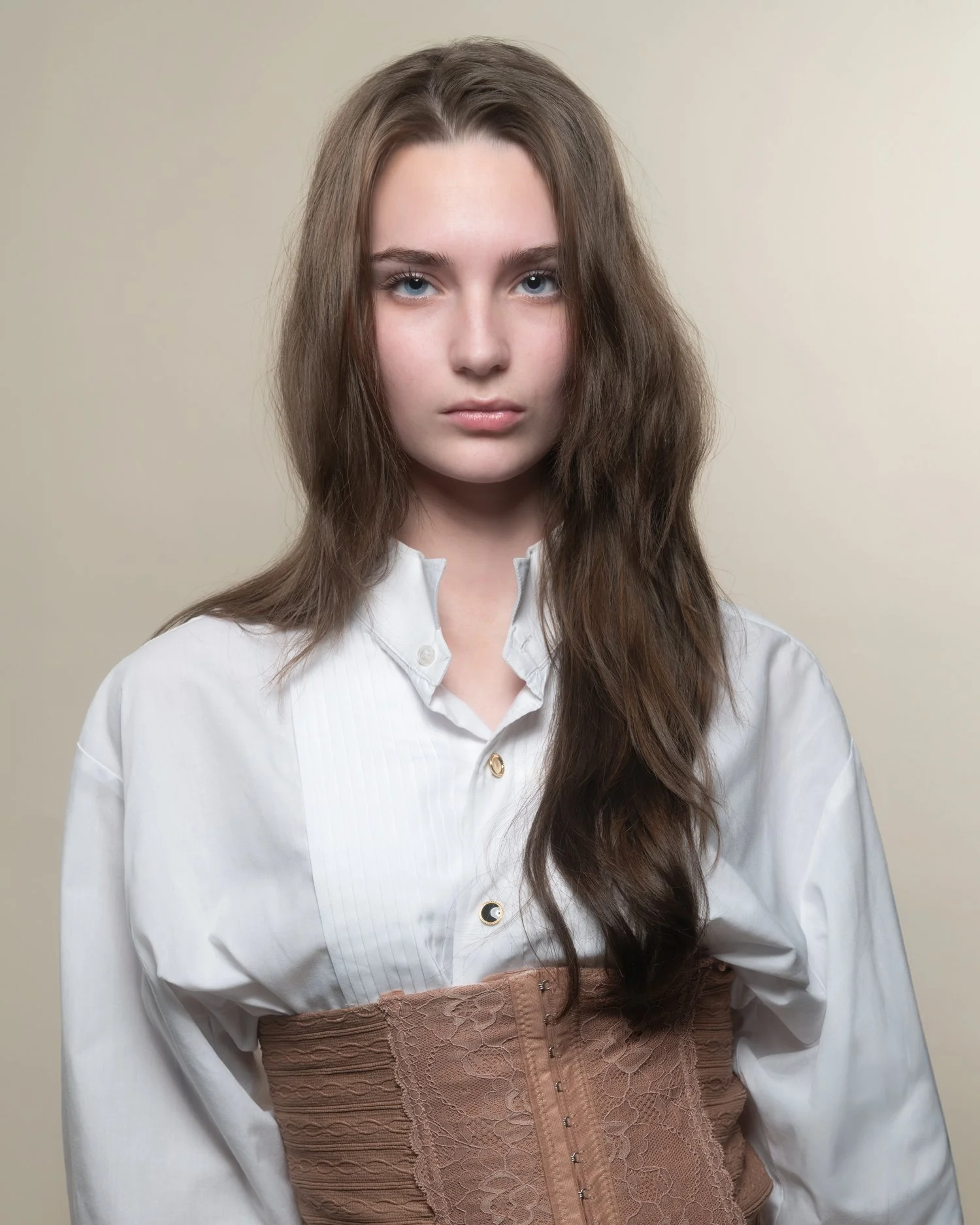 A young woman with long brown wavy hair and blue eyes wearing a white button-down shirt and a beige corset against a plain beige background.