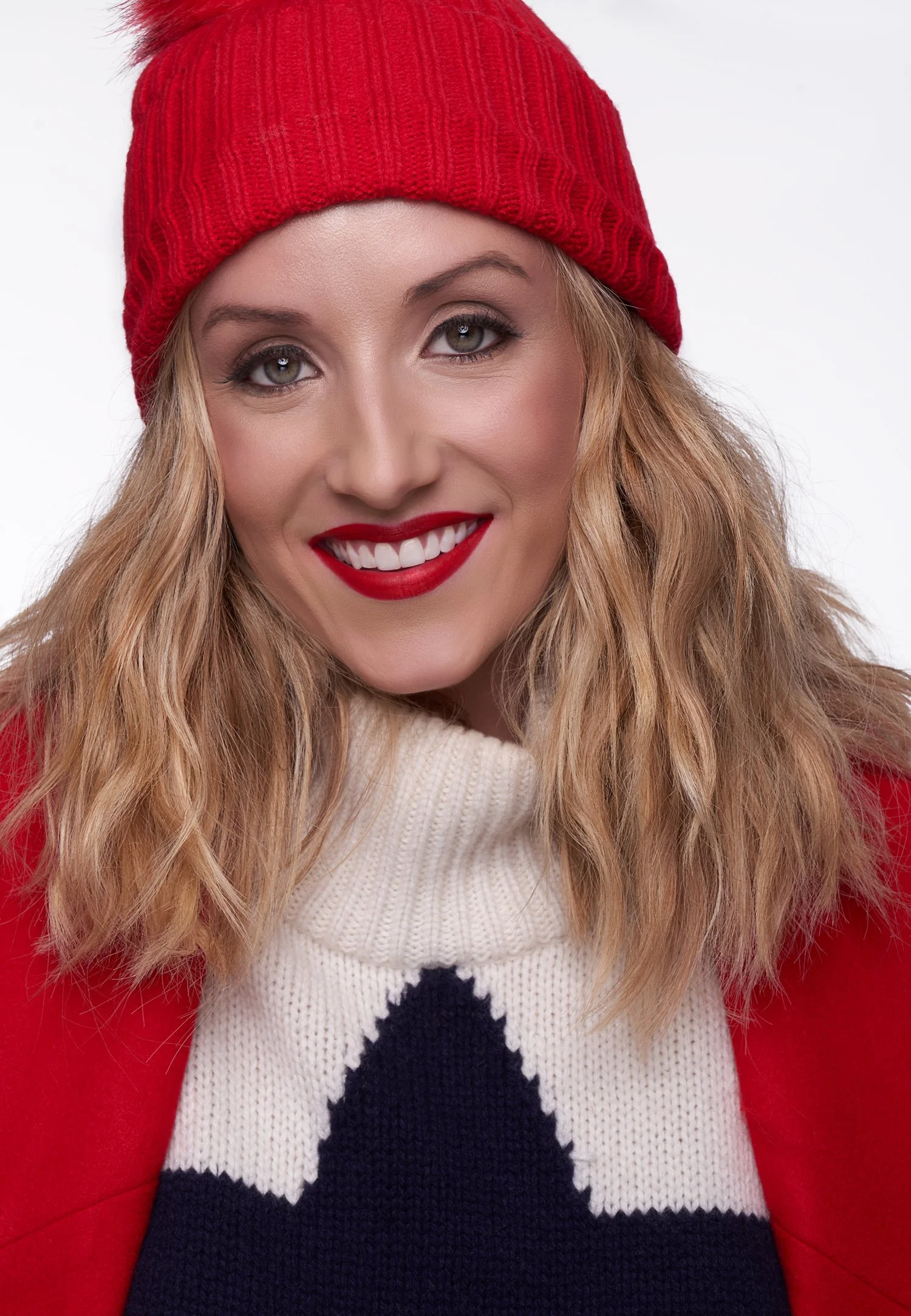 A woman with long wavy blonde hair wearing a red knitted beanie hat, a beige sweater, and a red cardigan. She has bright red lipstick and is smiling, showing her teeth. The background is white.