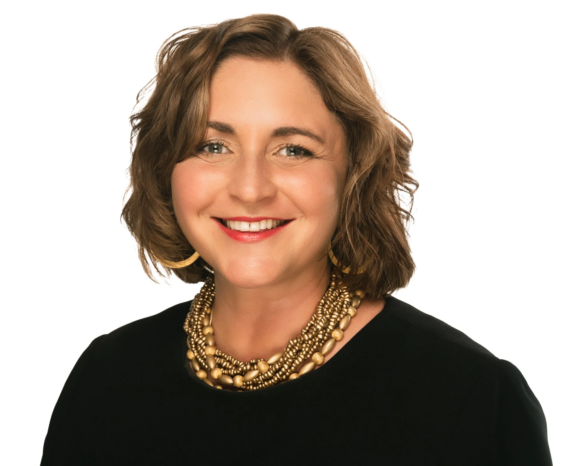 A woman with short, wavy, brown hair, wearing a black top, large gold earrings, and multiple layered gold necklaces, smiling against a white background.