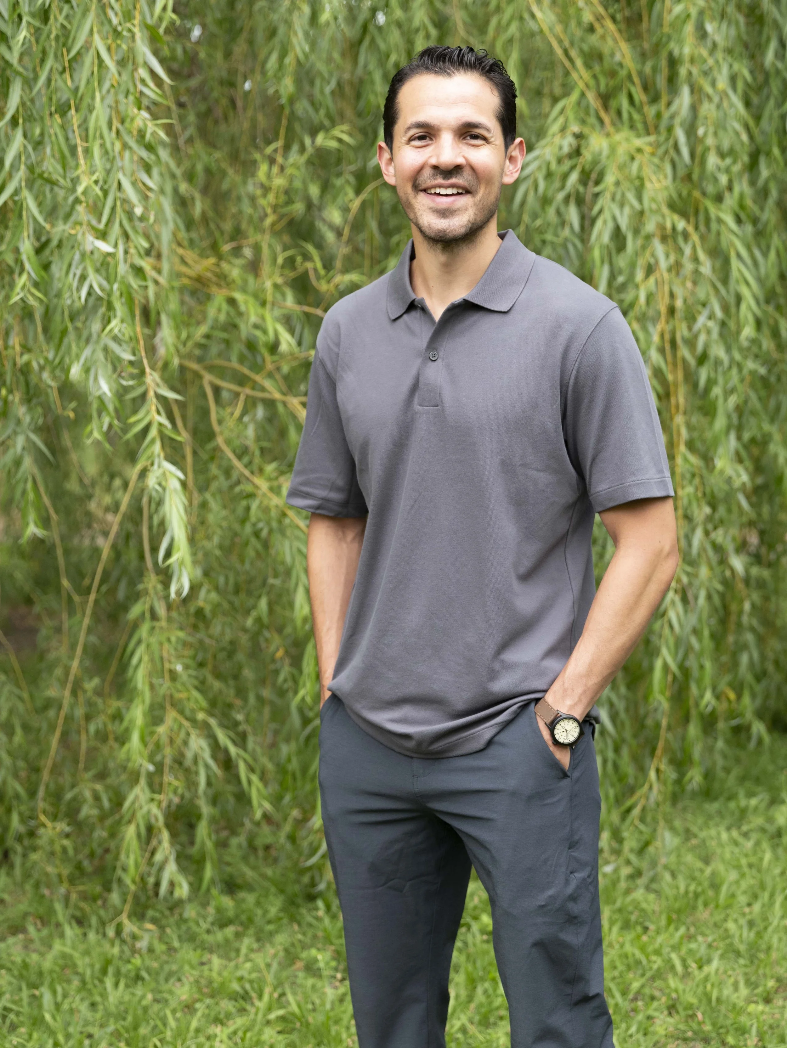 A man in a gray polo shirt and black pants standing outdoors near green trees, smiling with hands in pockets.