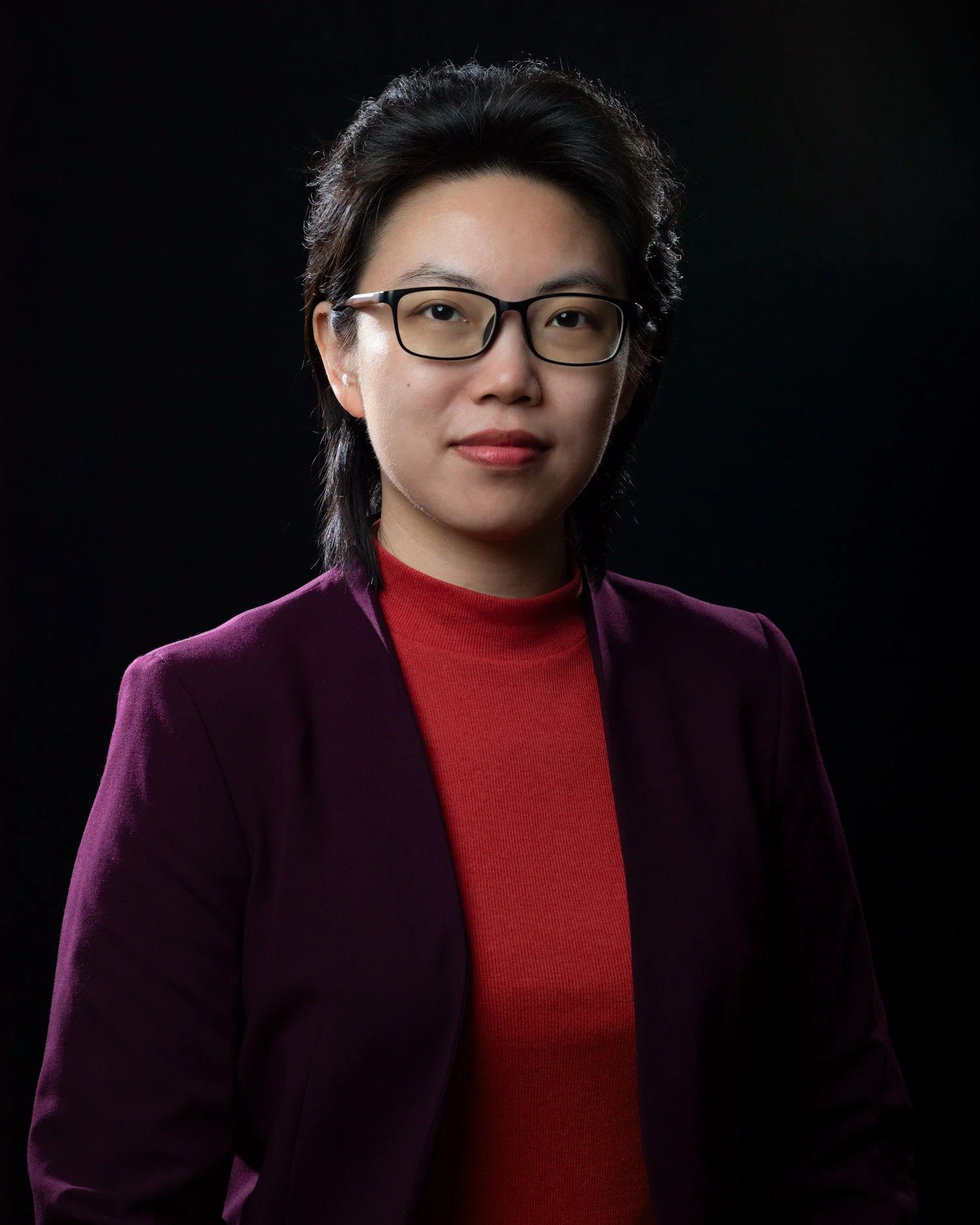Formal portrait of young Asian woman with medium-length black hair and glasses. She looks directly into the camera and wears a burgundy-color jacket over a bright red top against a solid black backdrop.
