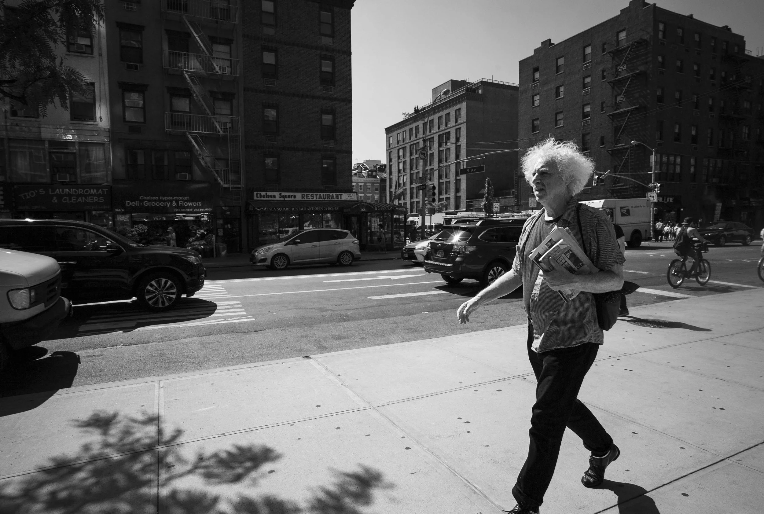 A black and white street scene with an elderly man with white, curly hair walking across the sidewalk while holding newspapers. Cars are parked along the street, and tall buildings are visible in the background. A cyclist is riding on the street.
