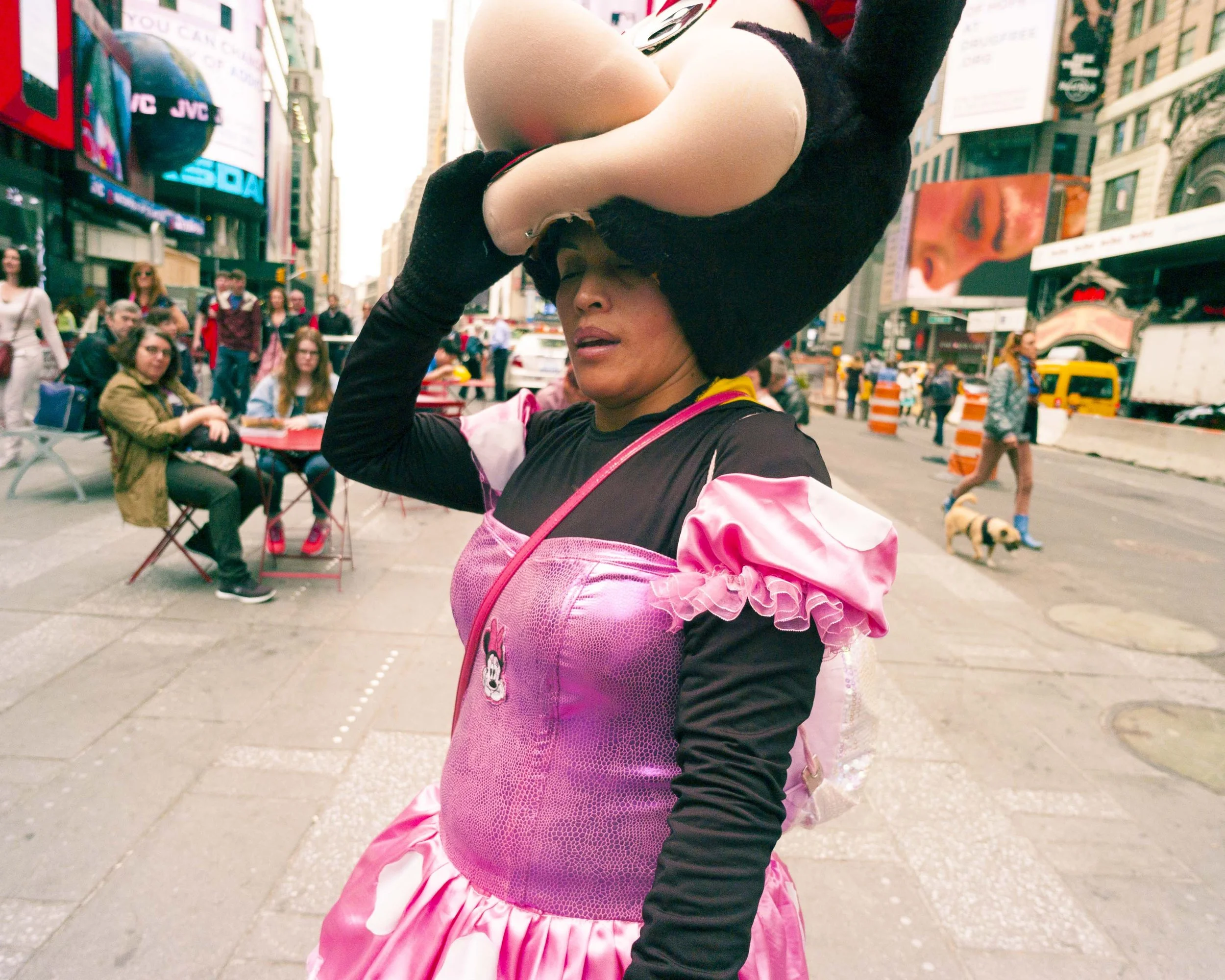 A woman dressed in a Minnie Mouse-themed costume on a city street, with a large pink bow and black gloves, surrounded by pedestrians and tall buildings.