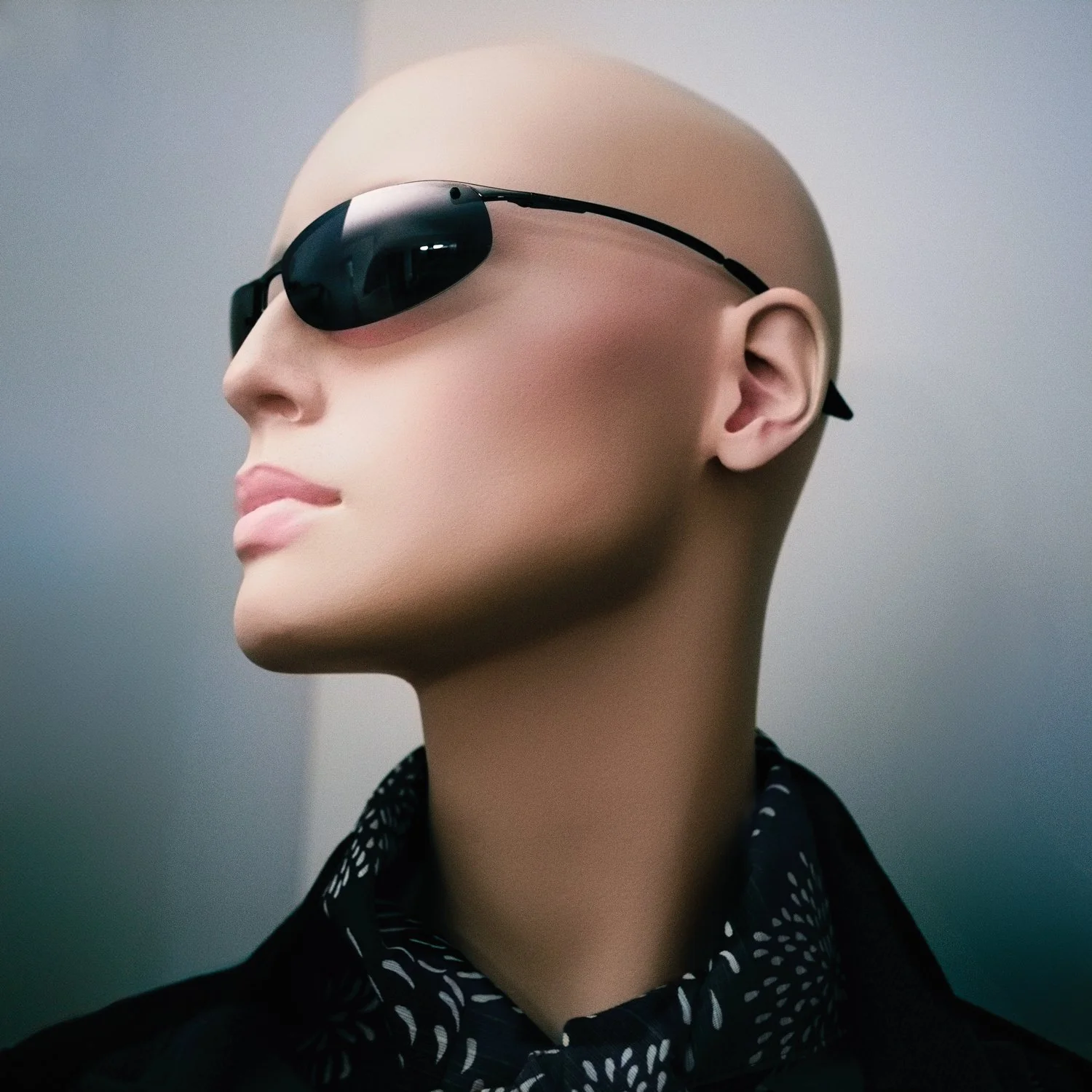 A mannequin with a bald head, wearing dark sunglasses, and dressed in a black jacket and patterned shirt.