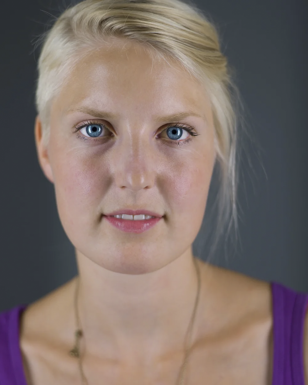 Close-up portrait of a young woman with blond hair, blue eyes, and fair skin, wearing a purple top and a thin necklace, against a dark background.