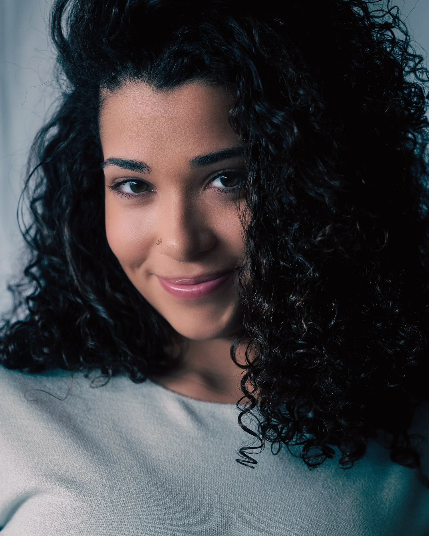 Closeup portrait of young female model with long, black curly hair and white top. She is looking directly into the camera with a closed-mouth slight smile, lit by diffused window light. 