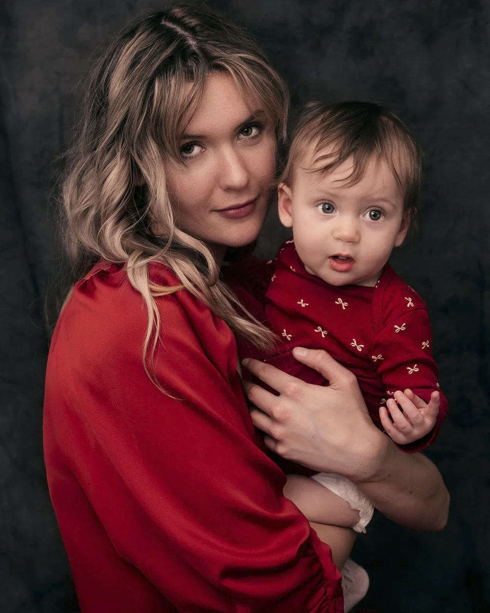 A woman with wavy blonde hair holding a baby girl with brown hair, both wearing red clothing, against a dark background.