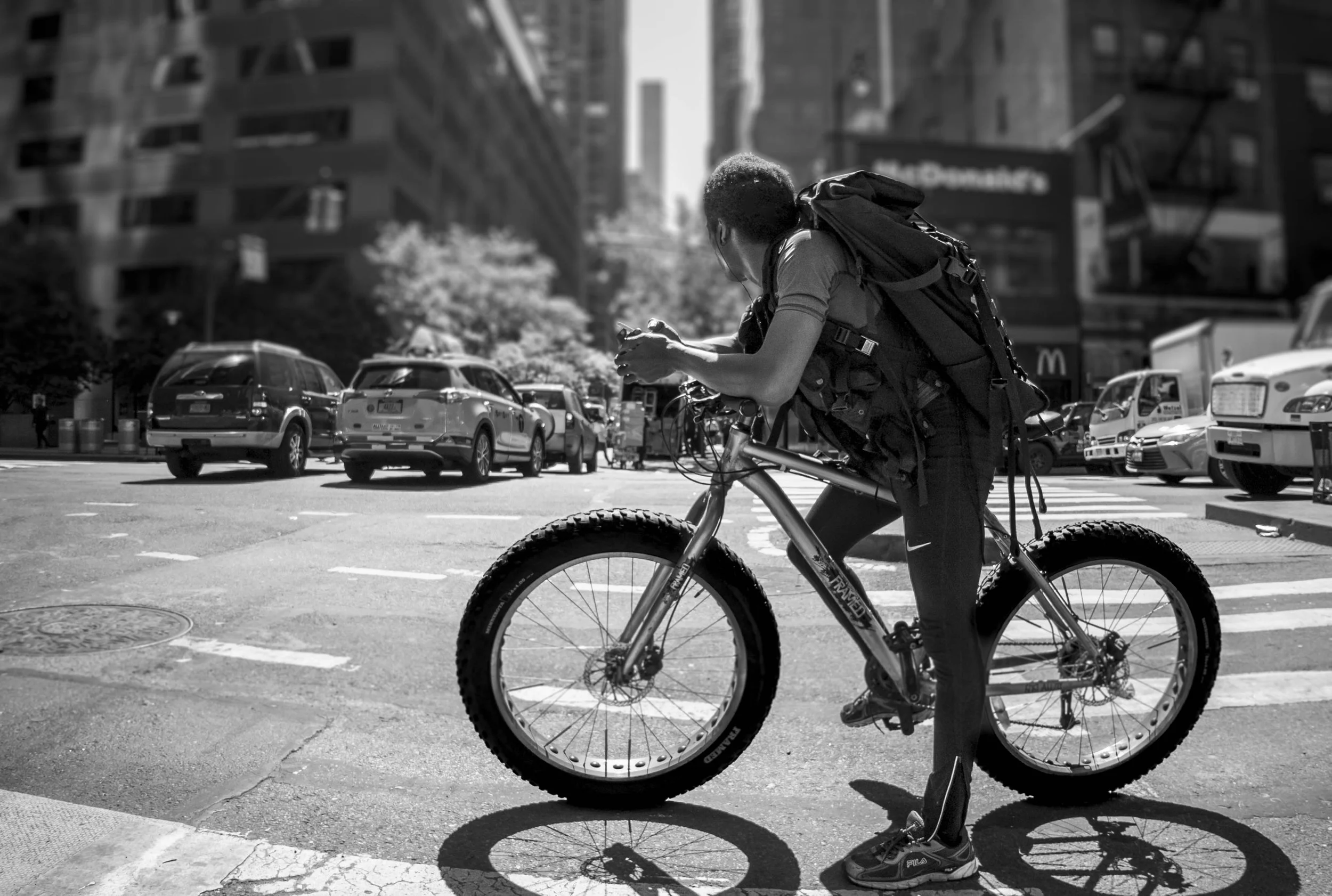 A person walking a bicycle across an urban intersection, looking at their phone, with cars and high-rise buildings in the background.