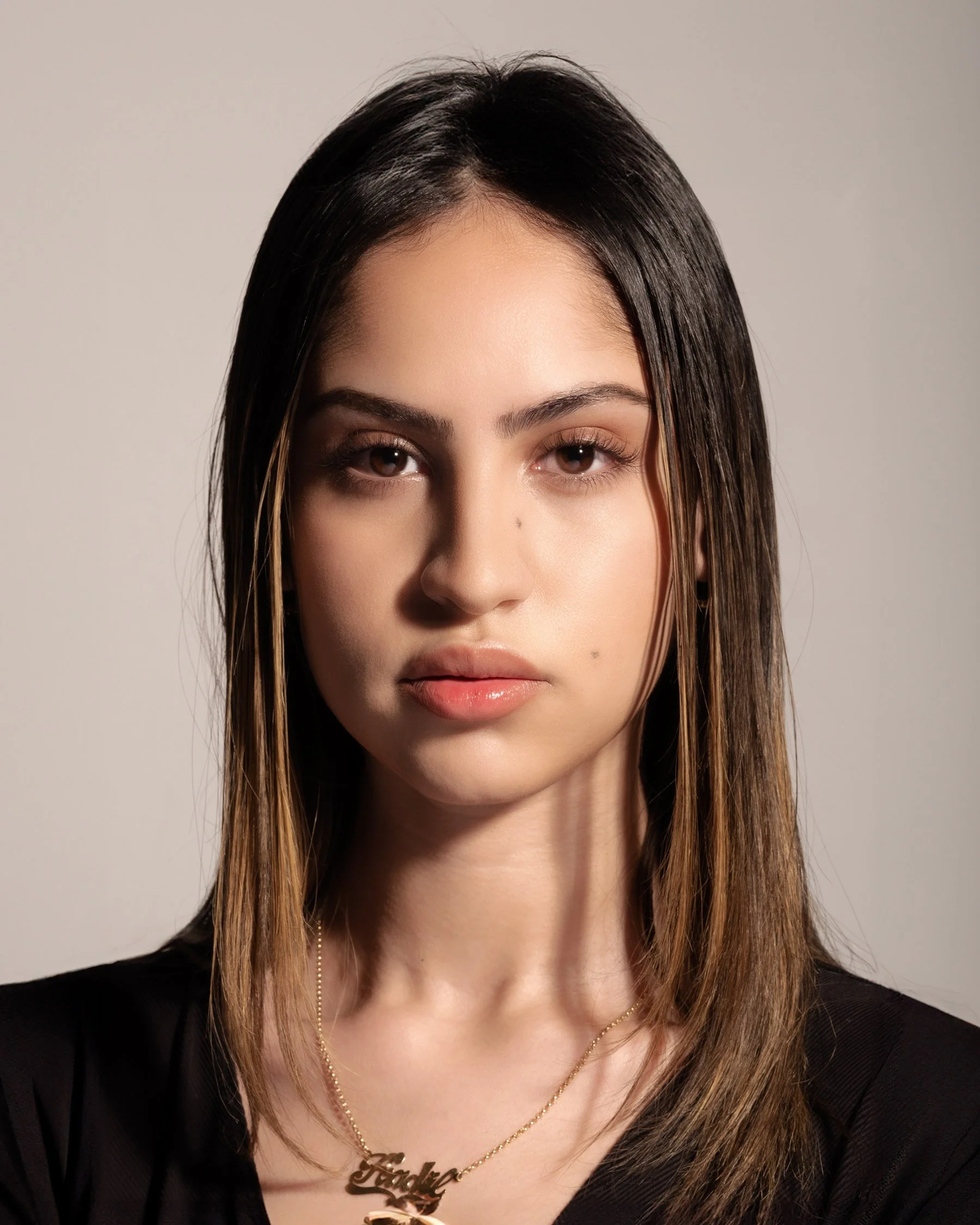 A young female model with a blank expression, stares into the camera. She has long, straight, dark hair with light brown highlights, large brown eyes, and full, pillowy lips and she wears a black v-neck top with a gold chain with a pendant that spell