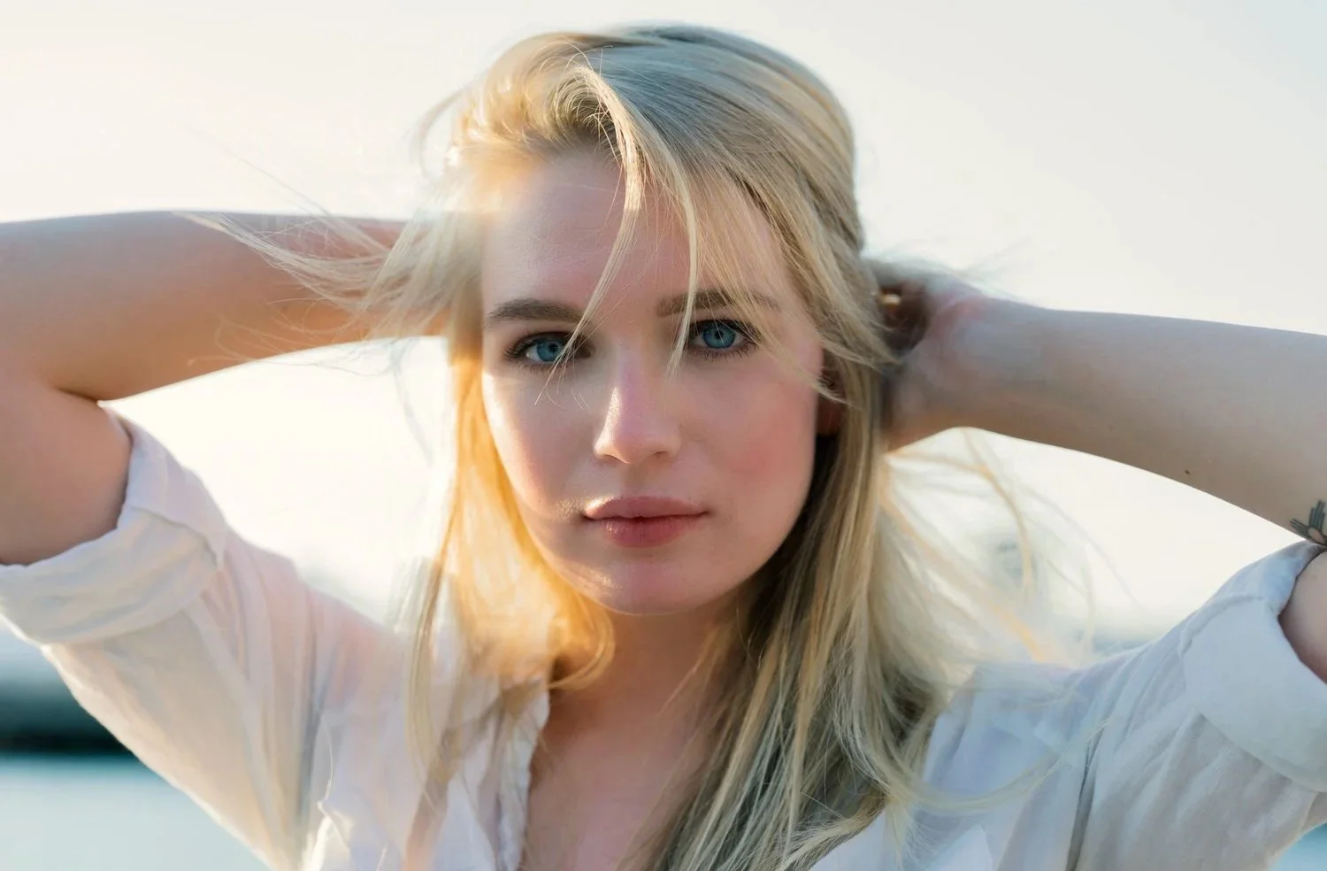 A young woman with blonde hair and blue eyes looking directly at the camera with hands behind her head outdoors during sunset.