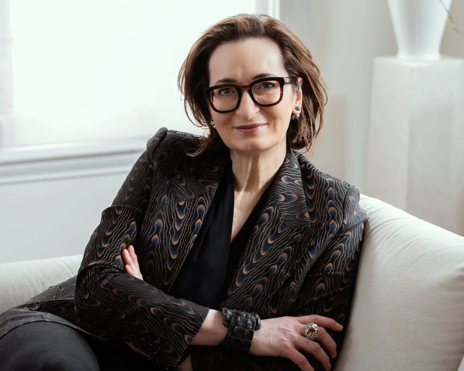 Headshot/portrait of a woman with medium-length brown hair and dark-rimmed glasses, seated on a sofa, bathed in natural window light.