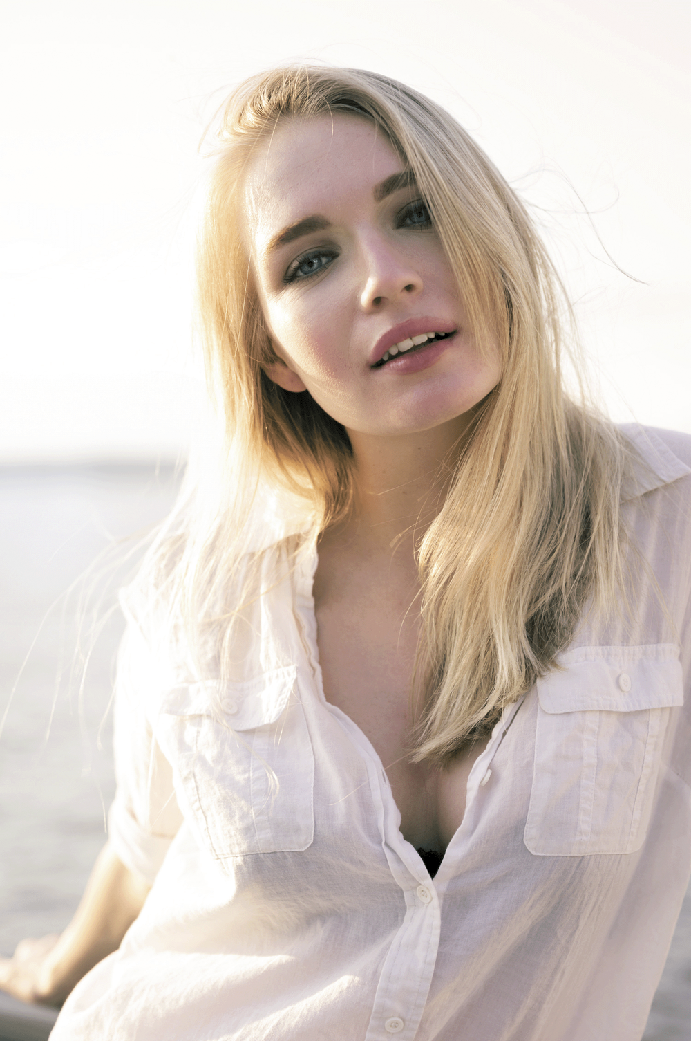 Close-up of a woman with long blonde hair, blue eyes, and an open white shirt, outdoors on a sunny day.