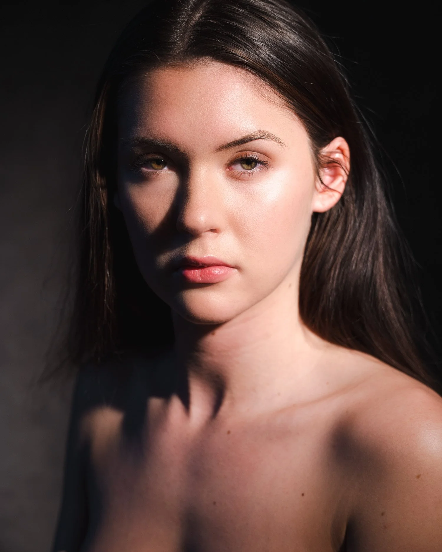 Close-up portrait of a young woman with long dark hair, looking directly at the camera with a neutral expression, contrasting lighting highlighting her facial features against a dark background.