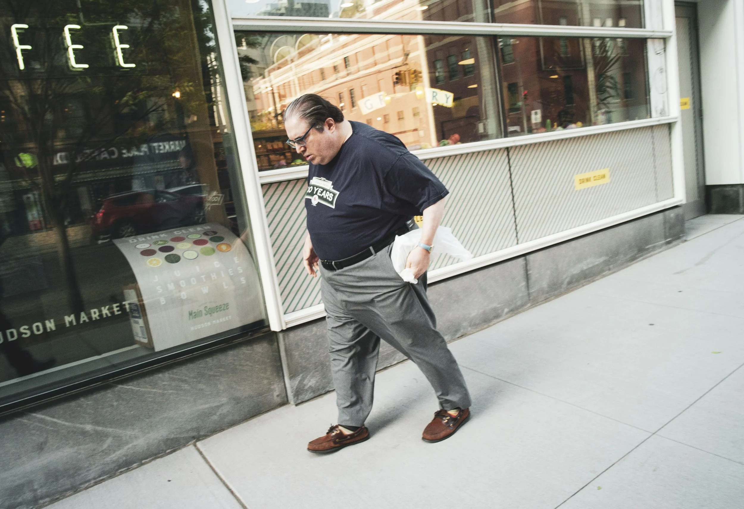 A man with glasses, wearing a black t-shirt and gray pants, walking on a sidewalk outside a storefront, carrying a white bag, with his head bowed and looking down.