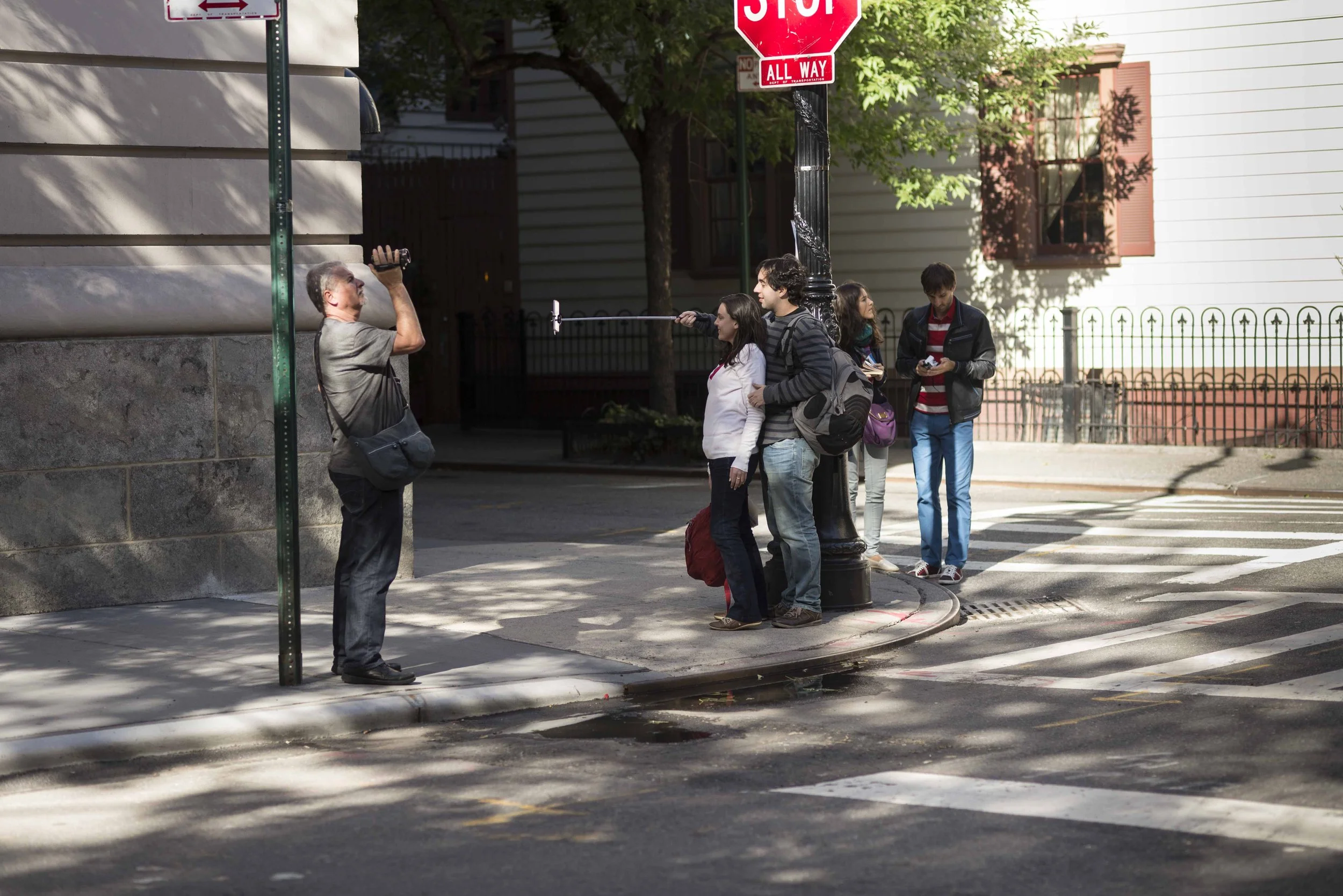 A group of young people standing at a street corner with a stop sign, a black lamppost, and a man taking a photo with a selfie stick. The background shows residential buildings and a tree with green leaves.