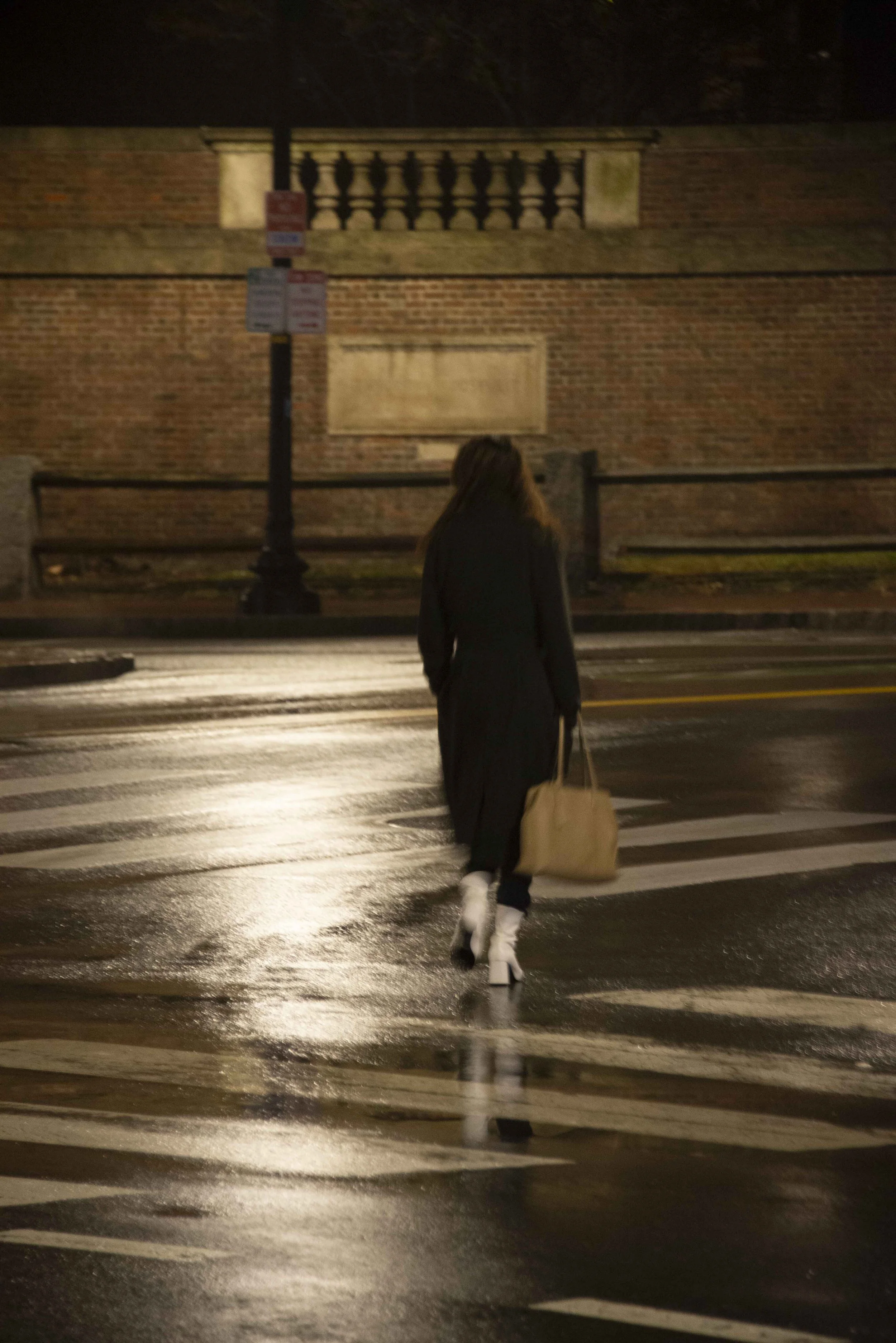 A person walking across a wet crosswalk at night. The person is carrying a large tan bag and has long hair. They are wearing a black coat, white high-heeled shoes, and white socks.