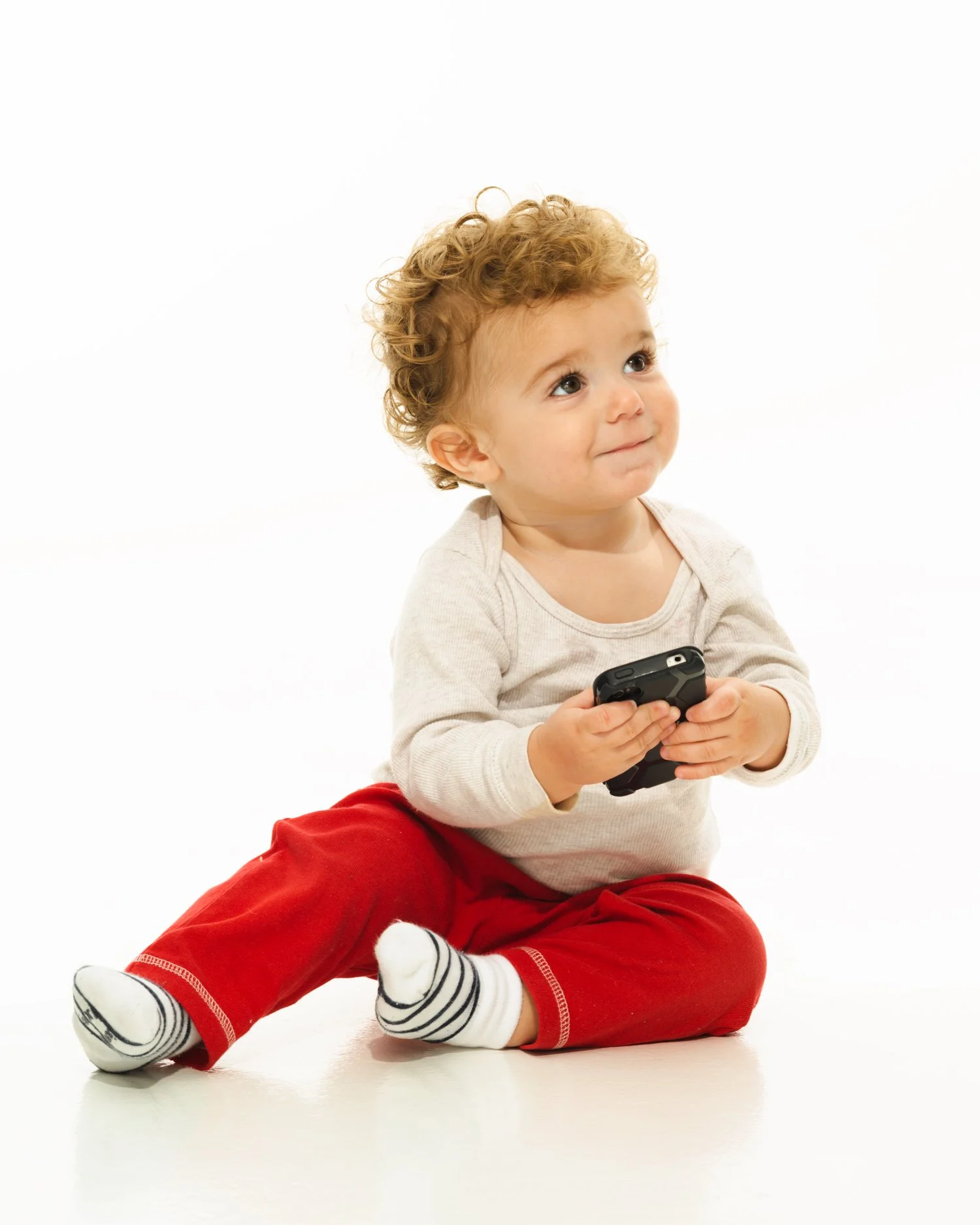 A toddler with curly blonde hair sitting on the floor against a plain white background. He is wearing a white henley shirt, red pants, with striped socks. He is holding a smartphone in his hands and looking slightly upward with a gentle smile.